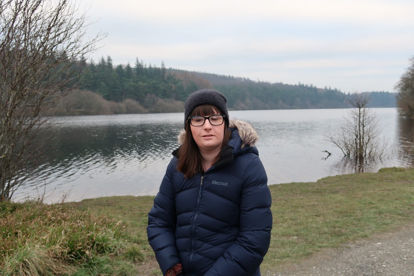 A photo of Holly wearing a coat and hat with a reservoir in the background