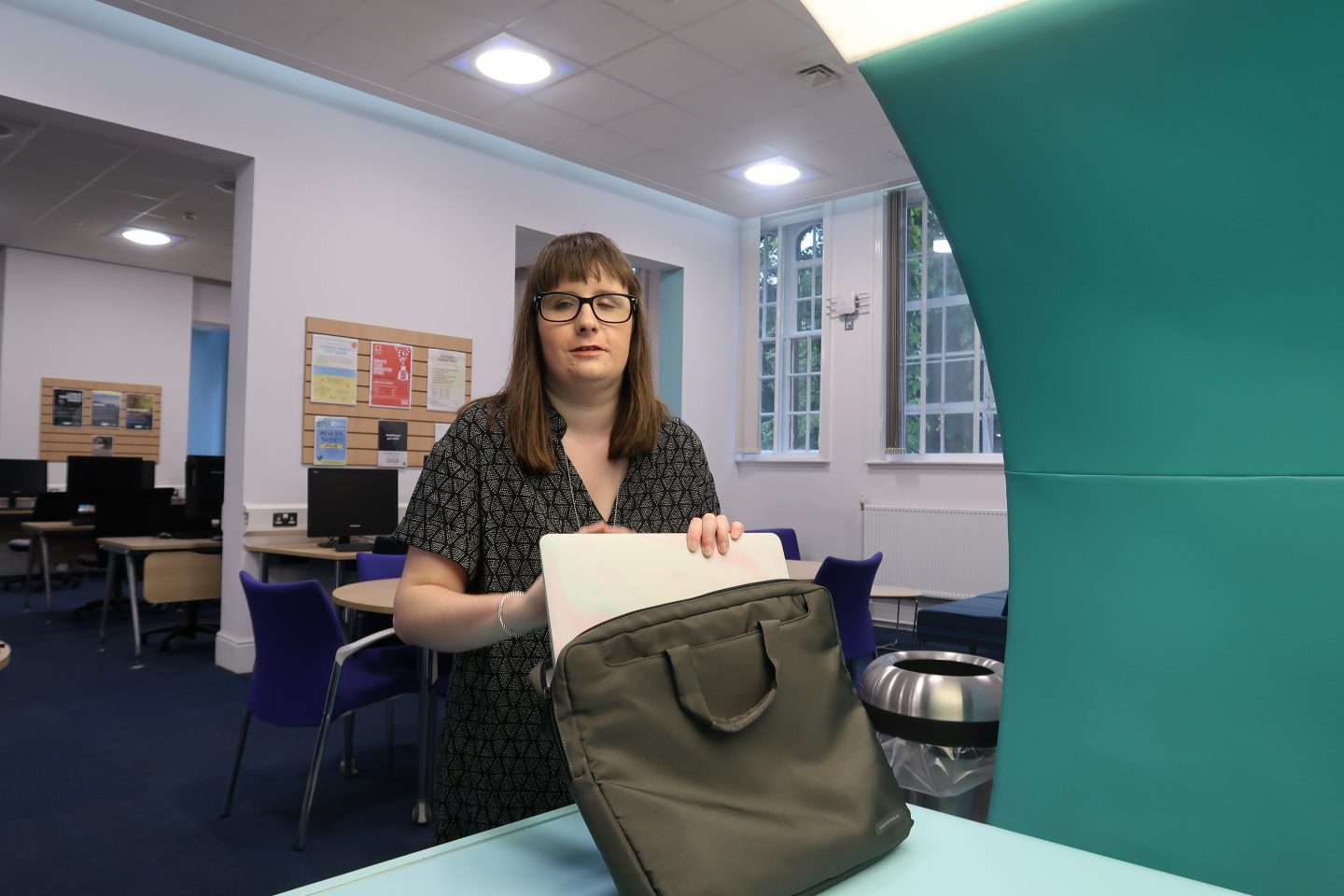 A photo of Holly stood up putting her laptop in her bag, there are chairs and tables in the background