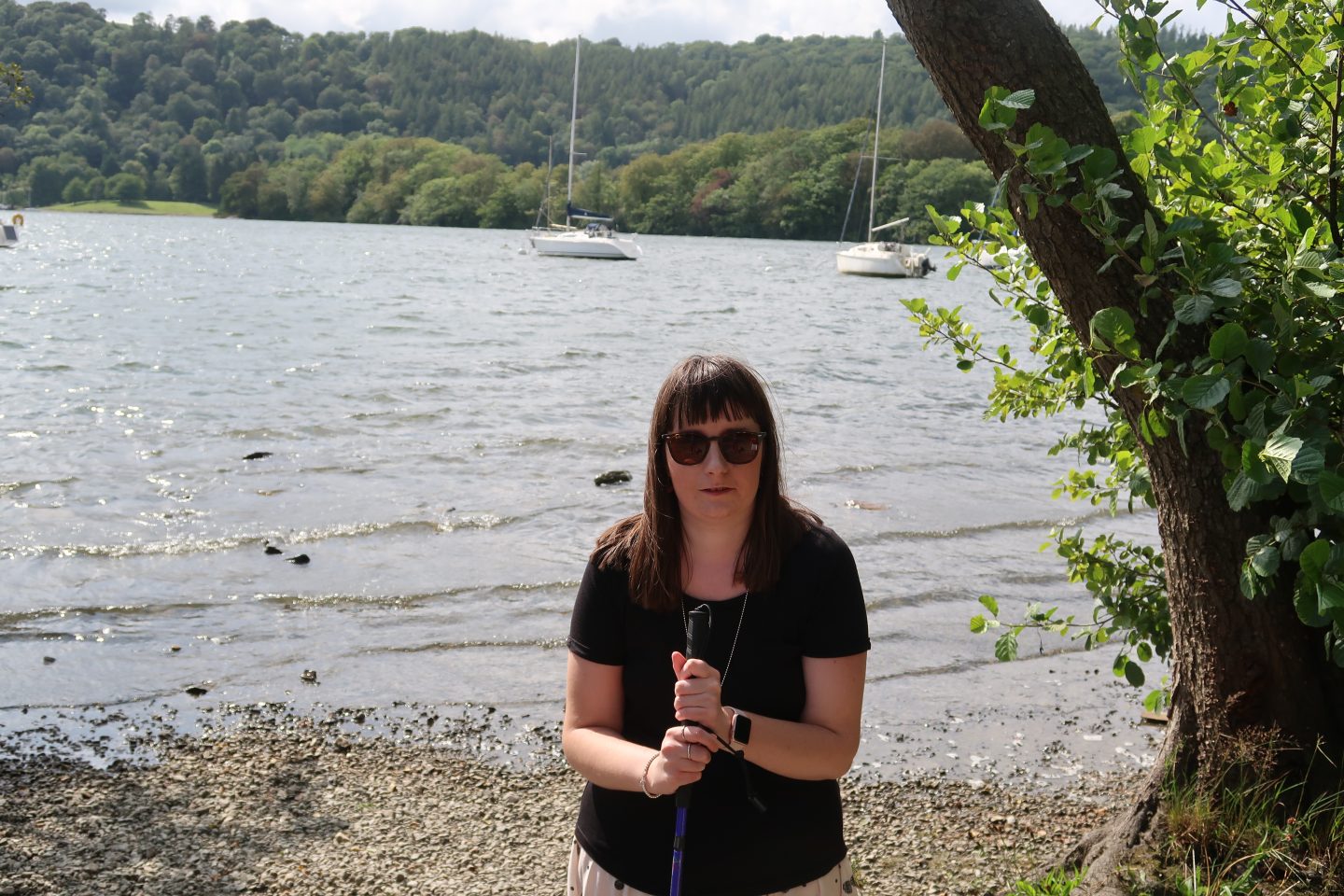 A photo of Holly holding her purple cane and waring sunglasses, half of the cane is visible in the photo, there are boats and a lake in the background