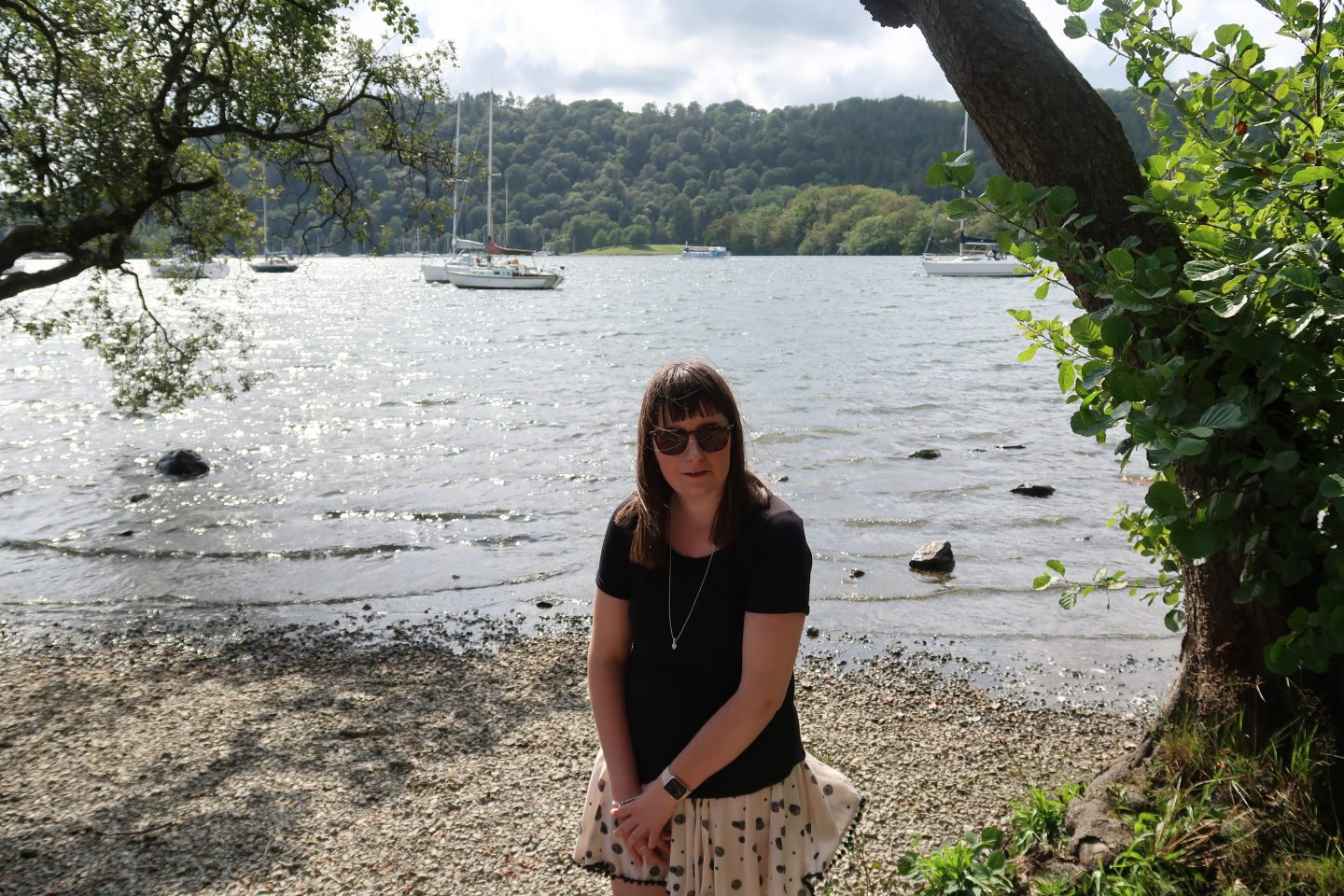 A photo of Holly stood in front of a lake, there are boats and trees in the background. She is wearing a black top and beige skirt