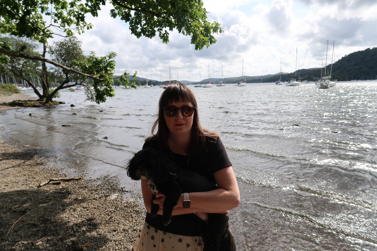 A photo of Holly cradling her small black dog who has white fur underneath her chin. There is a lake and boats in the background