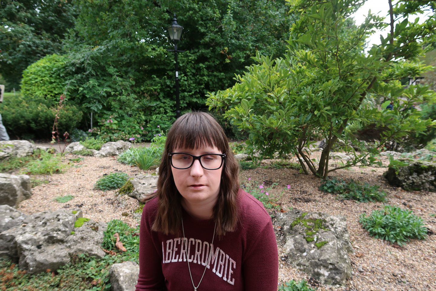 A close-up photo of Holly sat in front of a rockery which has plants and gravel around it, she is wearing a maroon Abercrombie & Fitch jumper