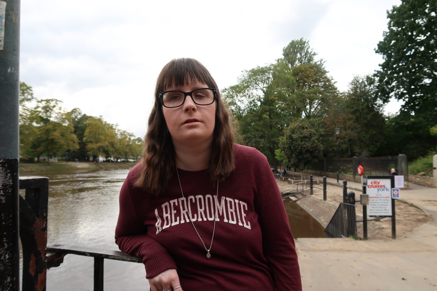 A close-up photo of Holly stood in front of a fence, there is a river, trees and footpath in the background, she is wearing a maroon Abercrombie & Fitch jumper