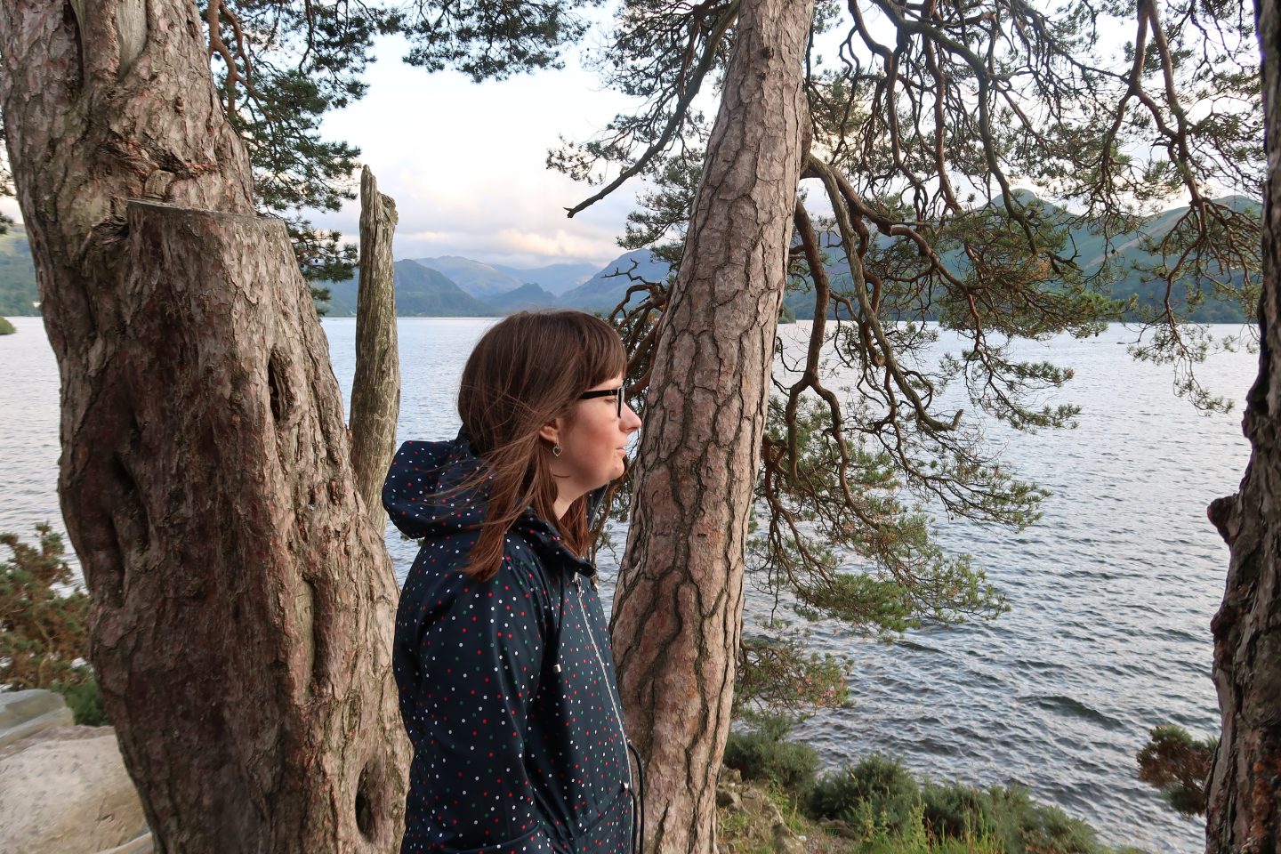 A photo of Holly stood sideways near a tree, she is looking towards a lake
