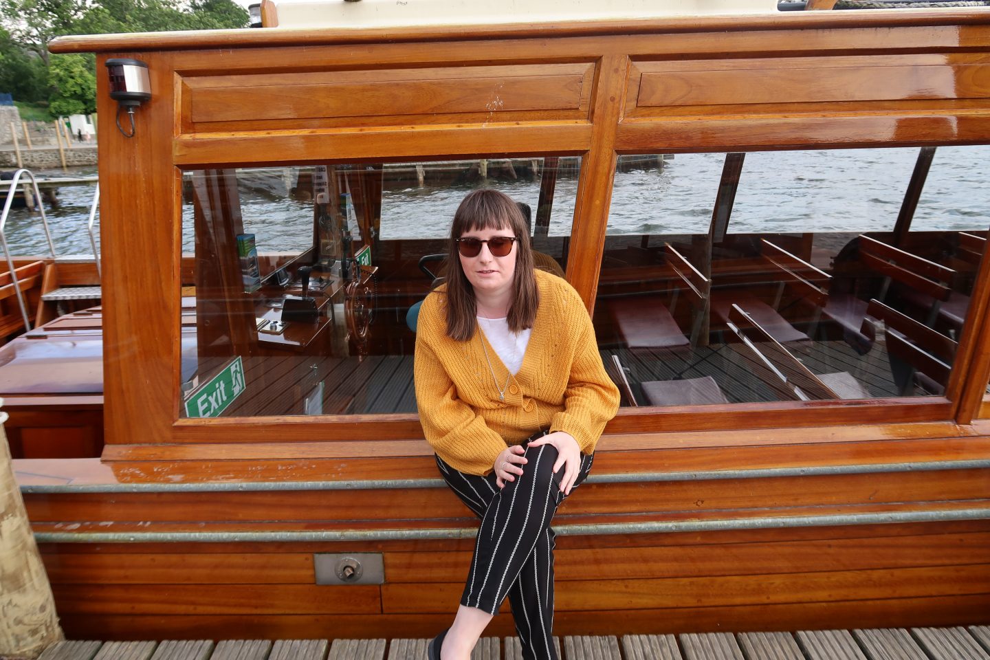 A photo of Holly sat on a boat, she is wearing a yellow cardigan and black striped trousers. There is a lake in the background