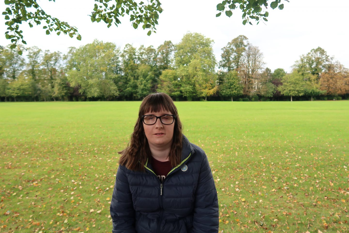 A photo of Holly wearing a blue coat, there is a green park behind her