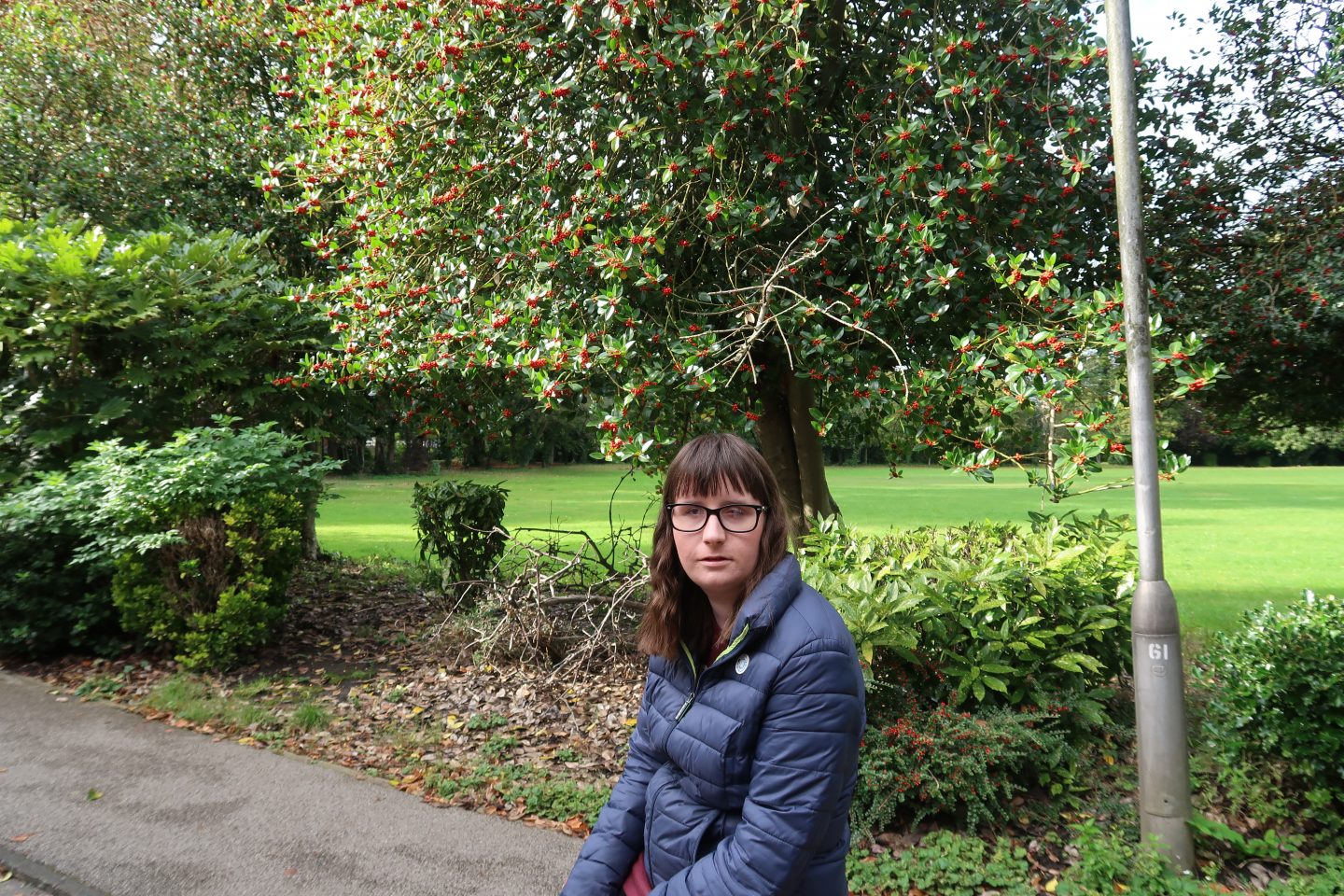 A close-up photo of holly sat on a large stone, she is wearing a blue Superdry jacket, there is a Holly tree which has red berries on in the background