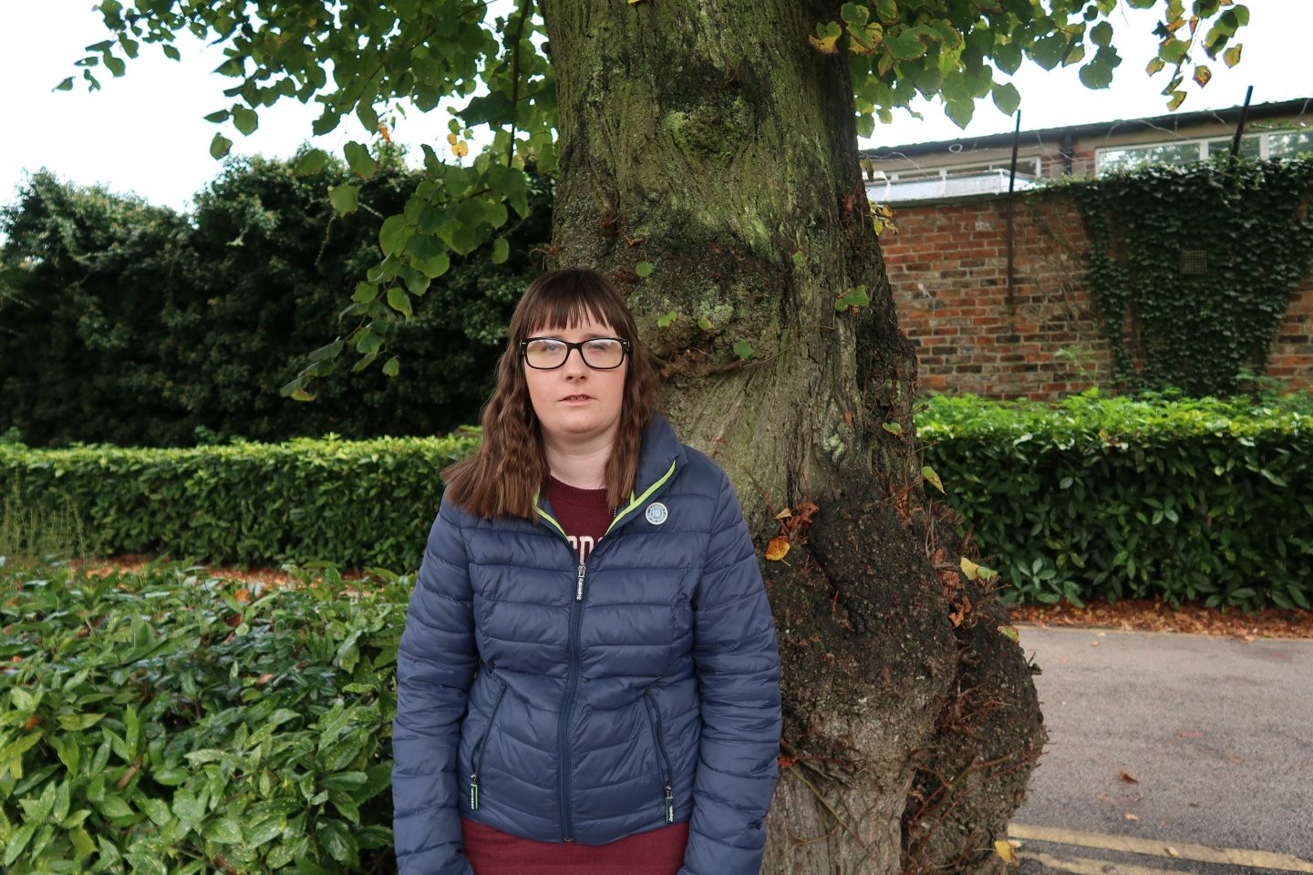 A photo of holly stood in front of a tree, she is wearing a blue superdry jacket and glasses