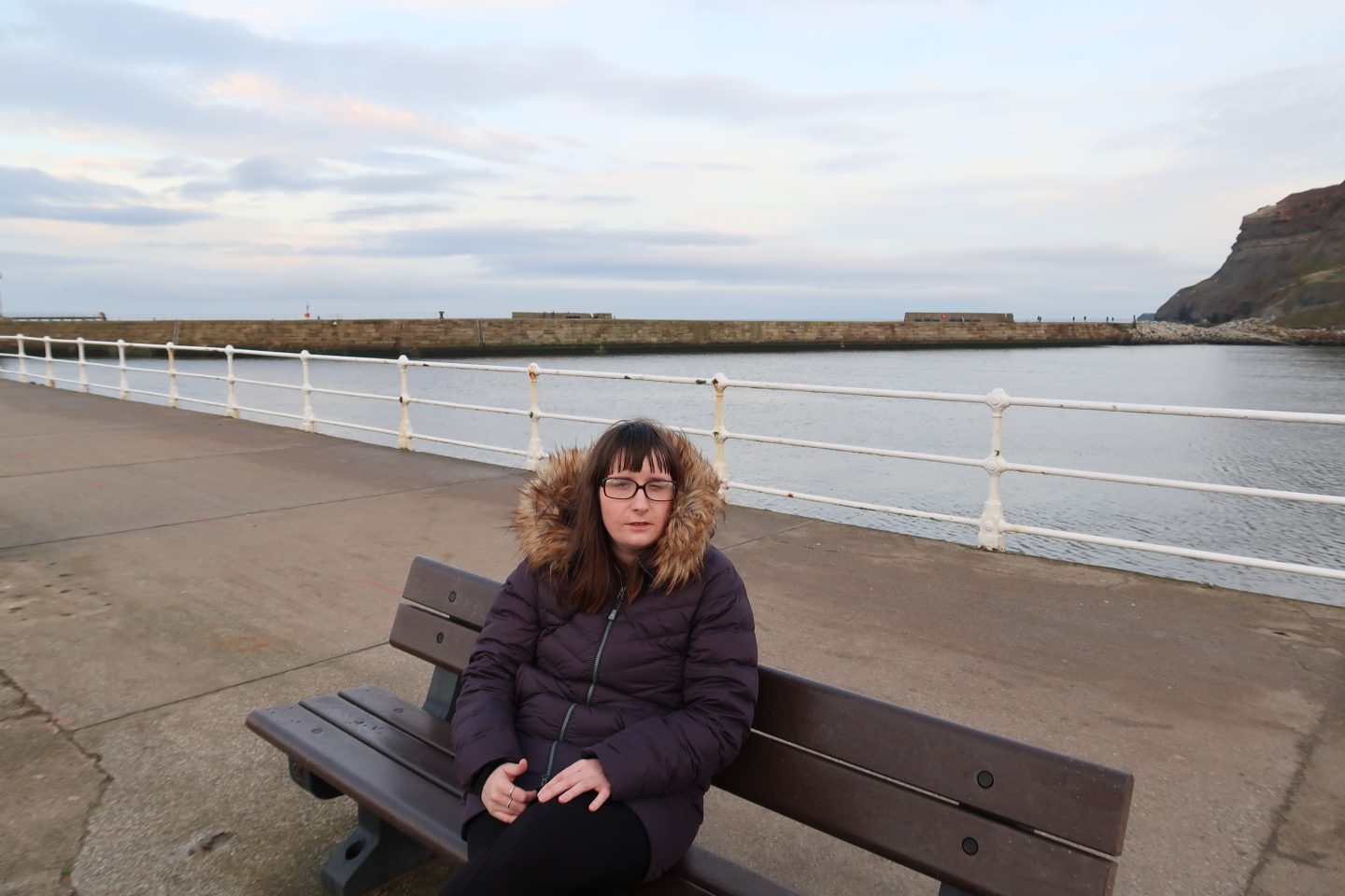 A photo of Holly sat on a bench, there is a harbour and cliffs in the background, she is waring a blue winter coat and black leggings