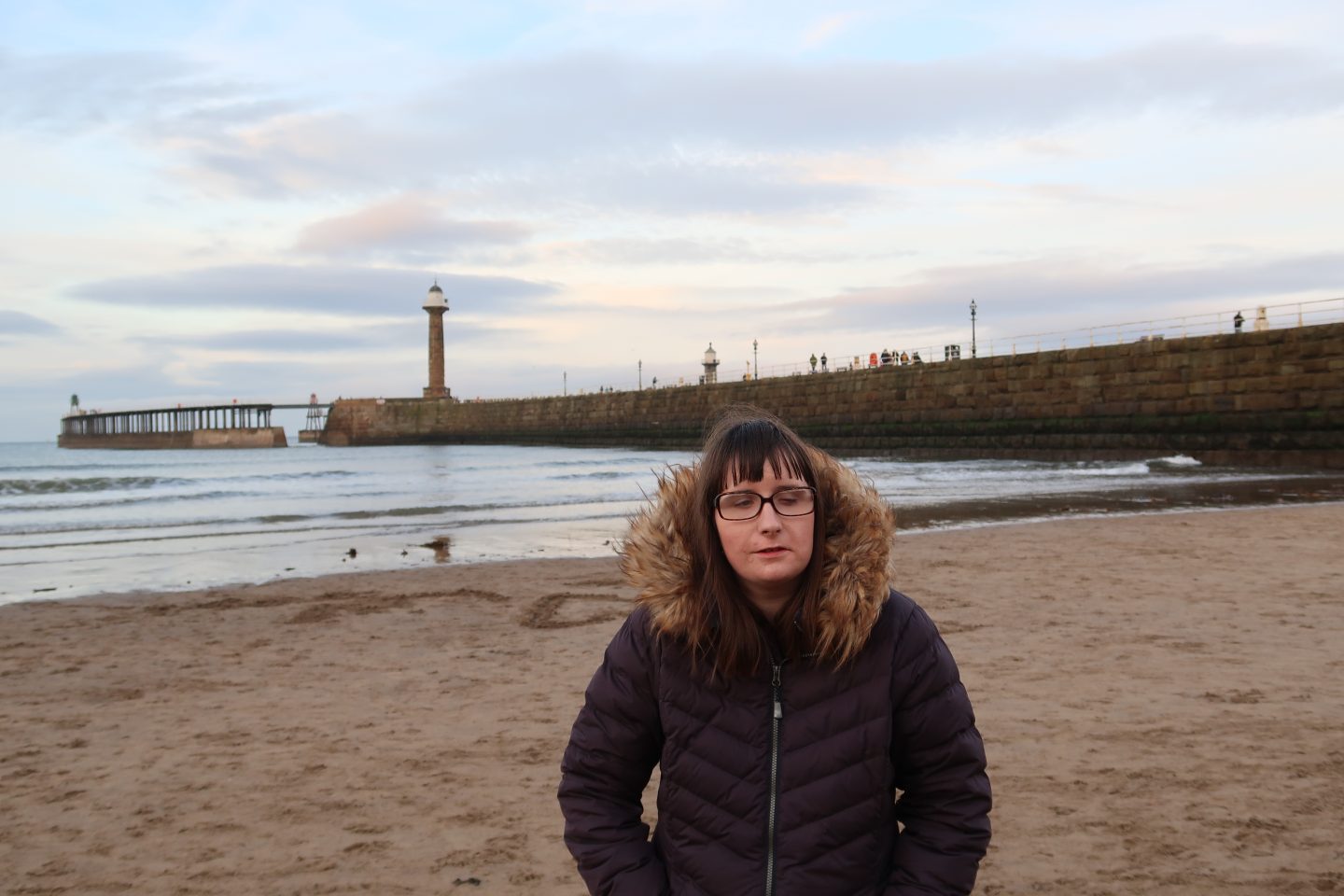 A photo of Holly on a beach, there is a pier and lighthouse in the background, the sea can be seen in the distance, she is wearing a winter coat and is stood with her hands in her pockets