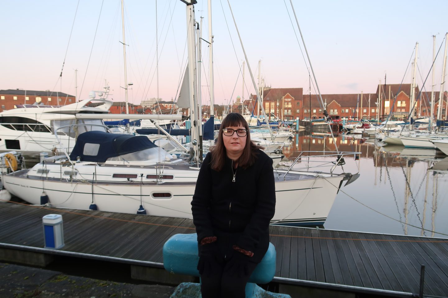 A photo of Holly sat on a blue stone, there are yachts behind and buildings in the background, she is wearing a black coat, black gloves and leggings