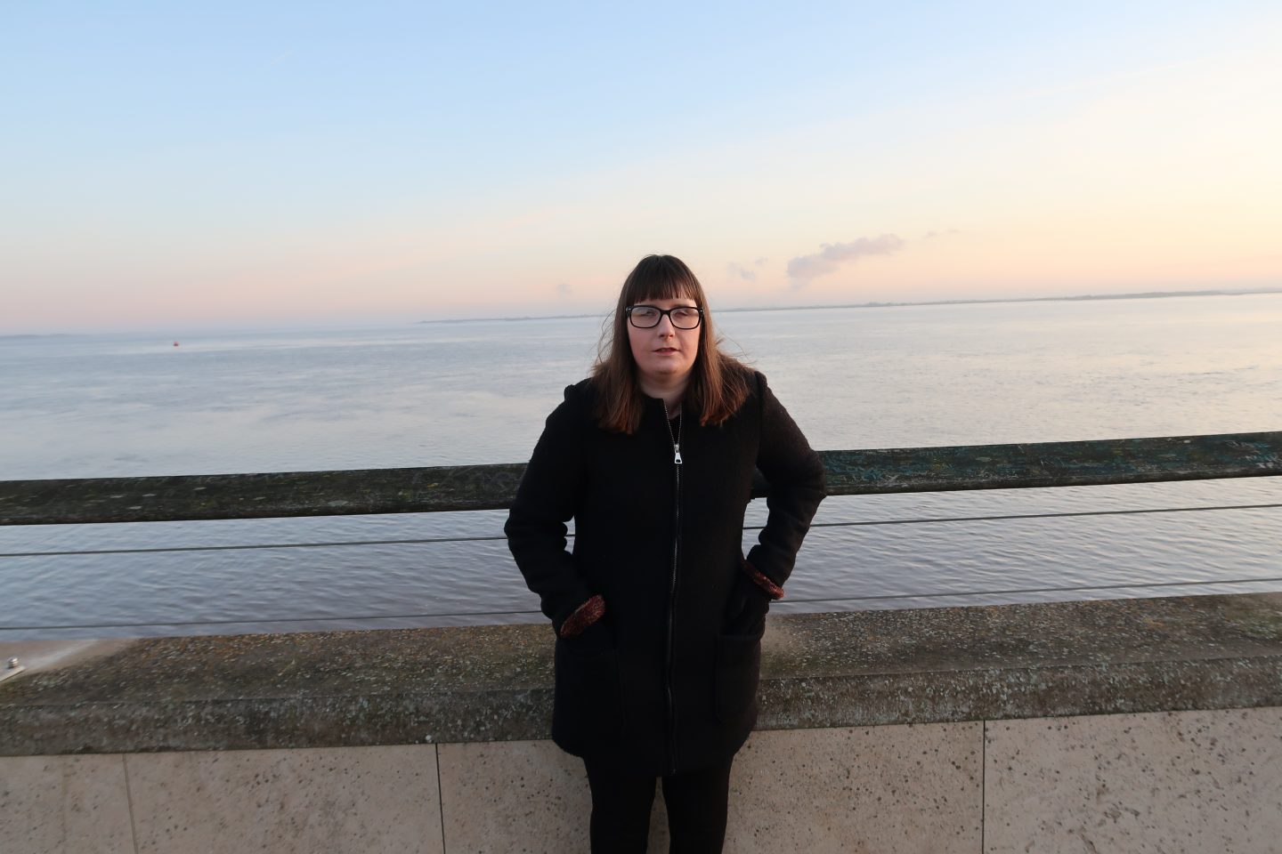 Holly stood in front of some railings with the sea behind her, she is wearing a black coat and leggings