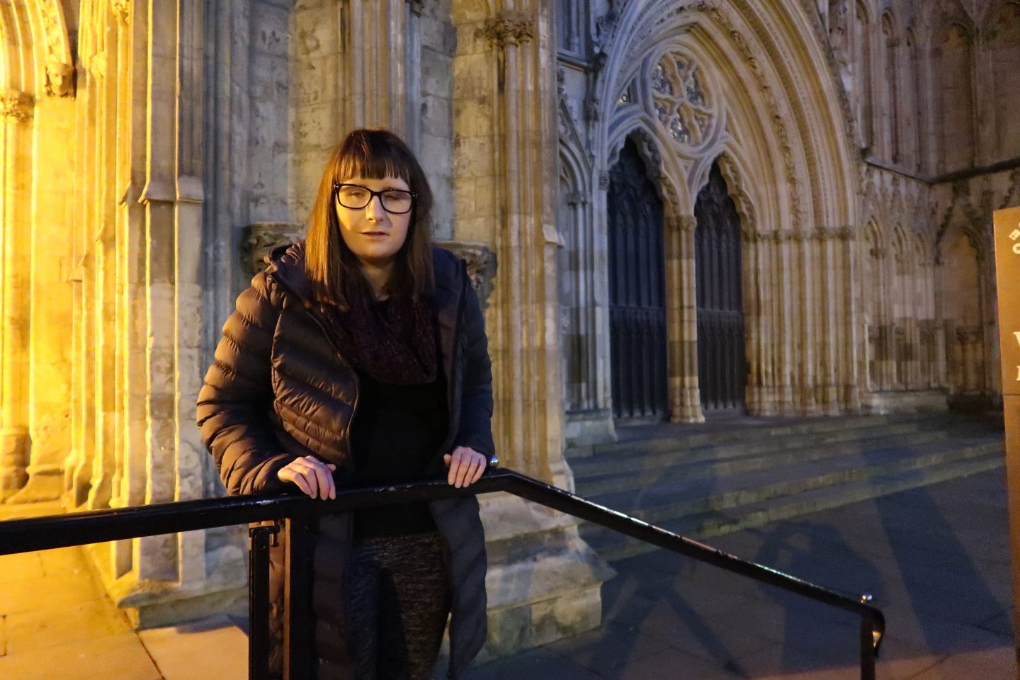 A photo of Holly stood on a step and leaning against a railing in front of York Minster, she is wearing a charcoal coloured coat, a burgundy scarf and black and grey trousers