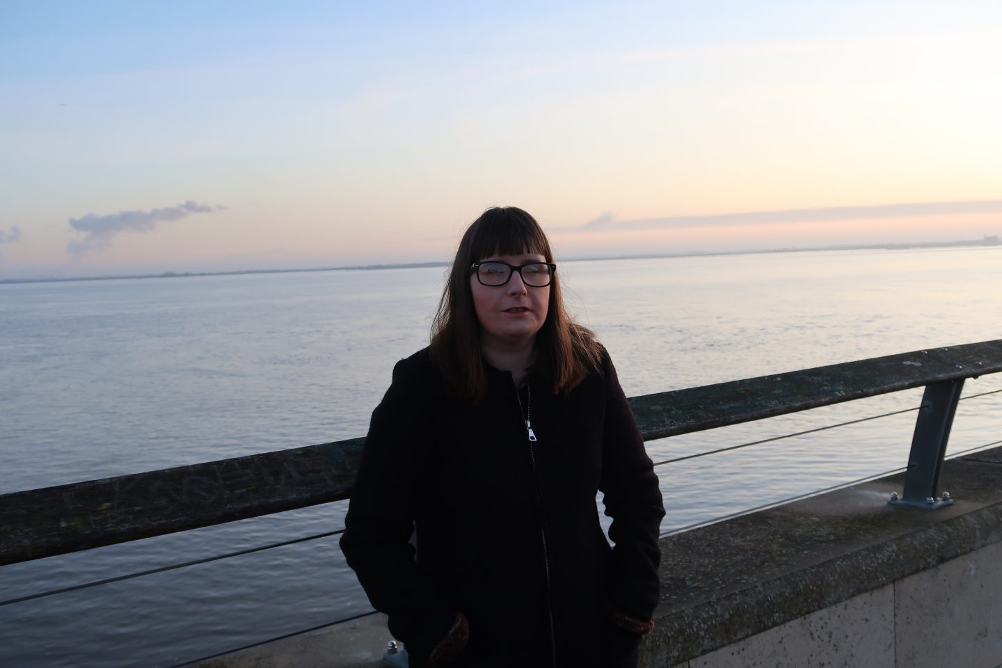 Holly stood against a railing with the sea behind her, she is wearing a black coat