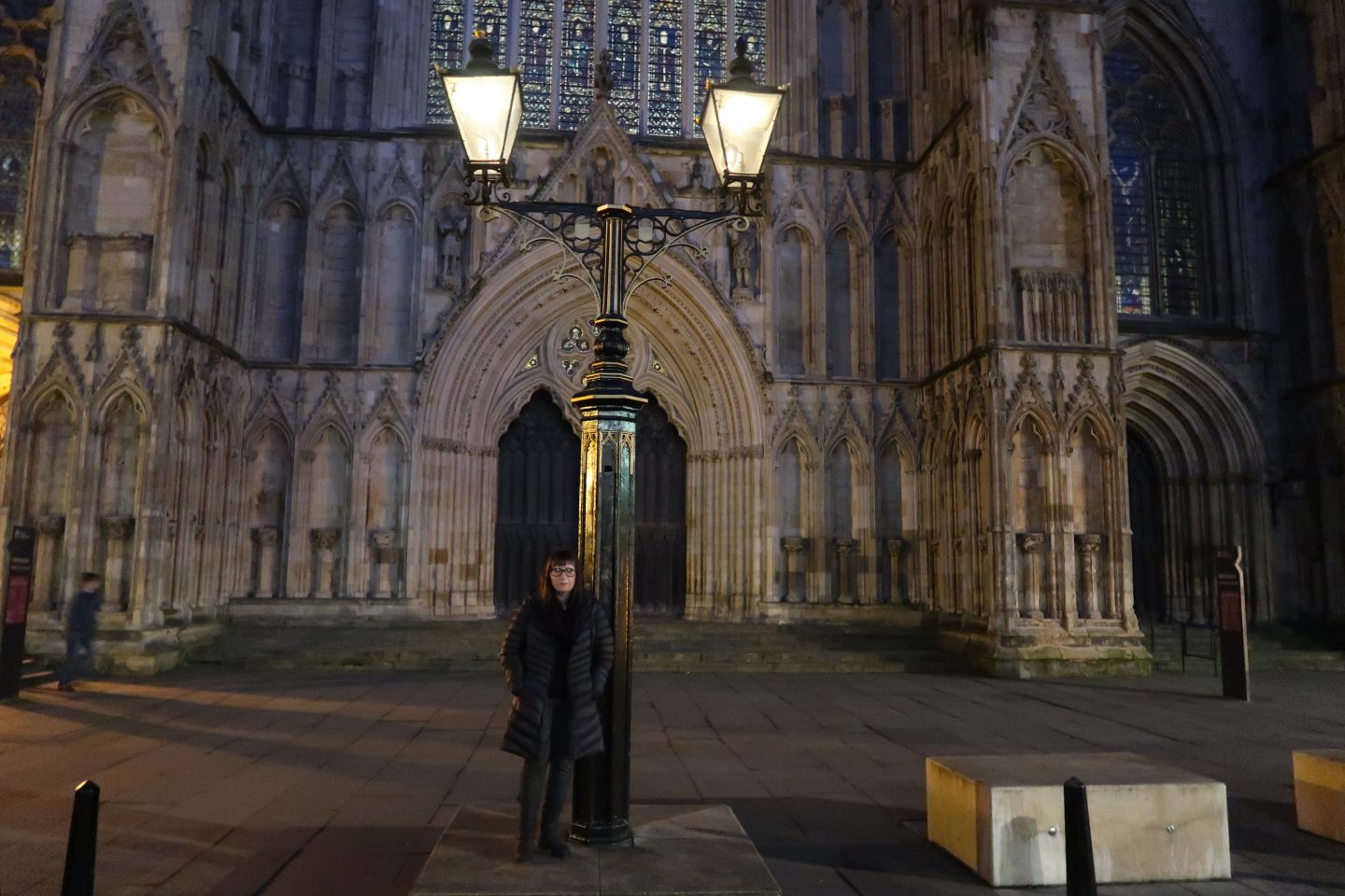 A photo of Holly stood in front of York Minster leaning against an old fashioned light, she is wearing a charcoal coloured coat, a burgundy scarf, black and grey trousers and grey boots
