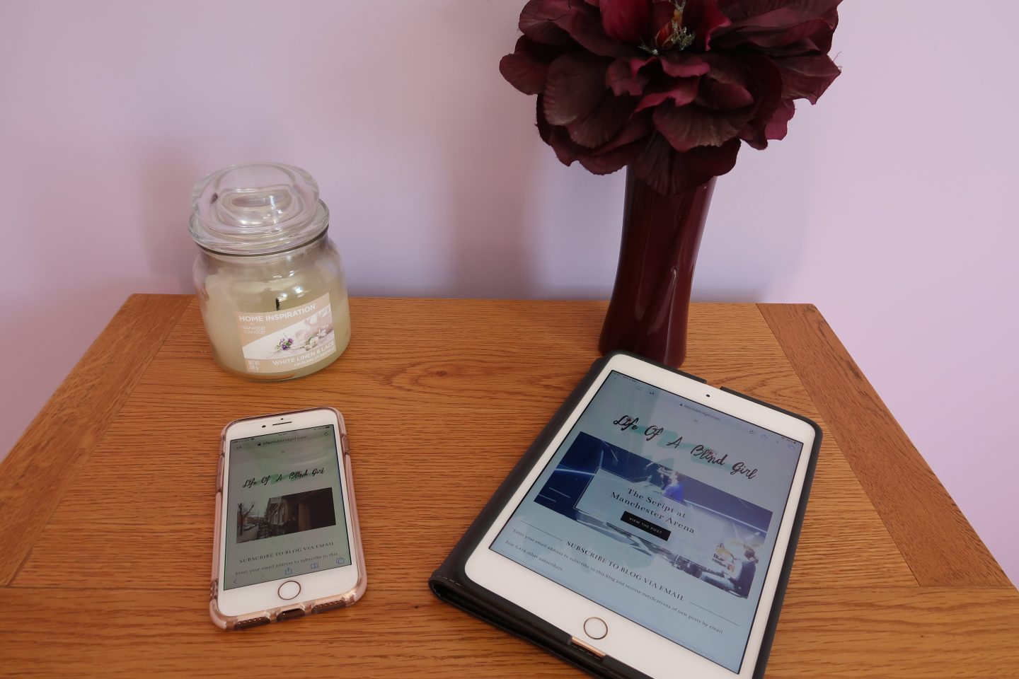 An iPhone and iPad on a table with Holly's blog posts showing on both screens, there is a candle and a purple flower in the background