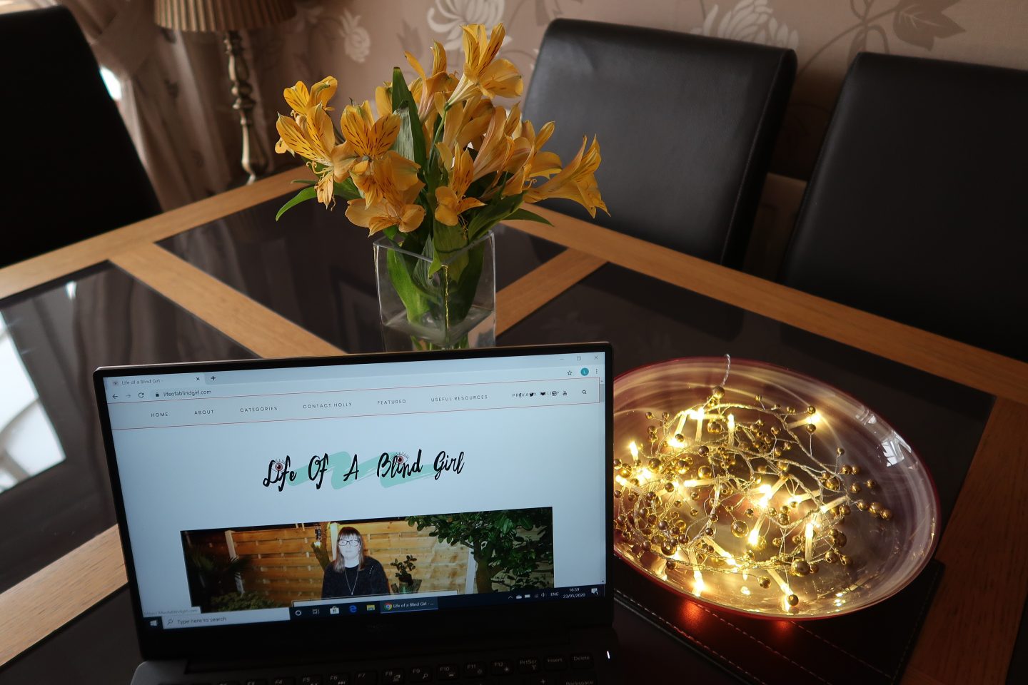 One of Holly's blog posts on a laptop screen, there is a red bowl that has lights in and a vase of yellow flowers behind it