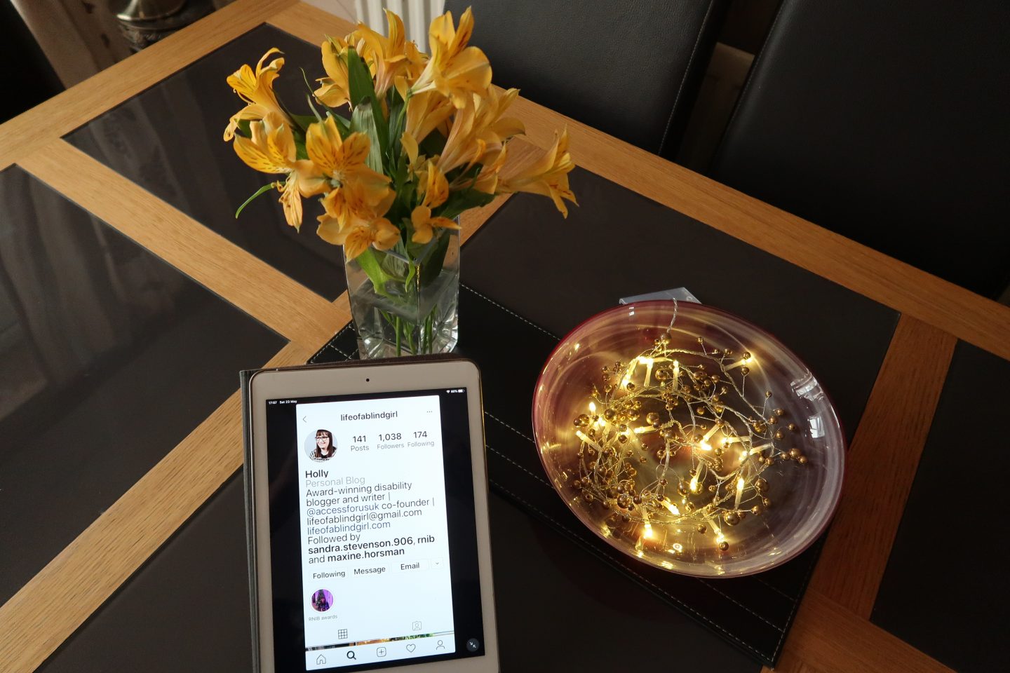 Photo taken from above showing Holly's Instagram profile on an iPad, there is a vase of yellow flowers and lights in a red bowl behind it