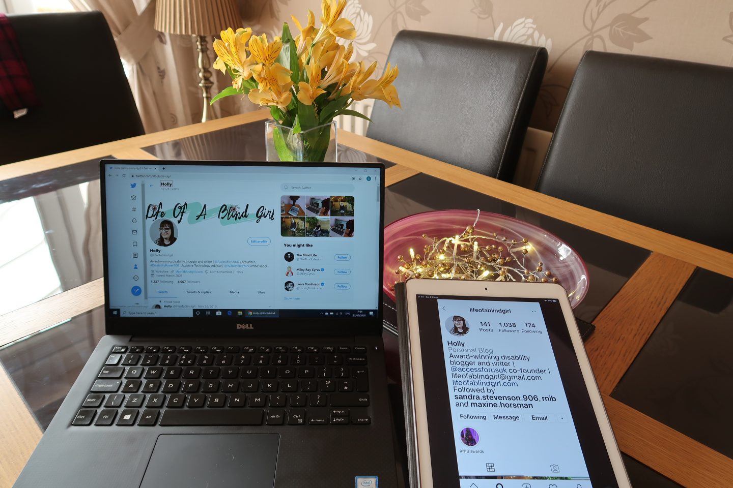 Holly's Twitter profile on a laptop screen and her Instagram profile on an iPad screen, there is a vase of yellow flowers and lights in a red bowl behind them