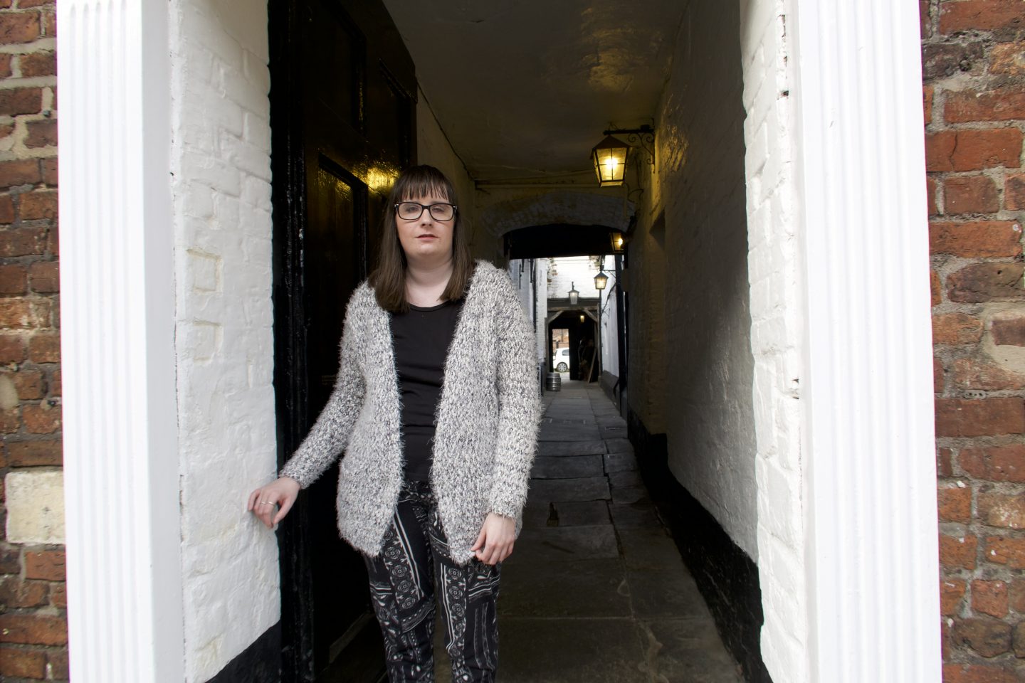 Holly stood in front of an alleyway, there are lamps on the wall behind her. She is wearing a black top, black and white trousers and a multi-coloured cardigan