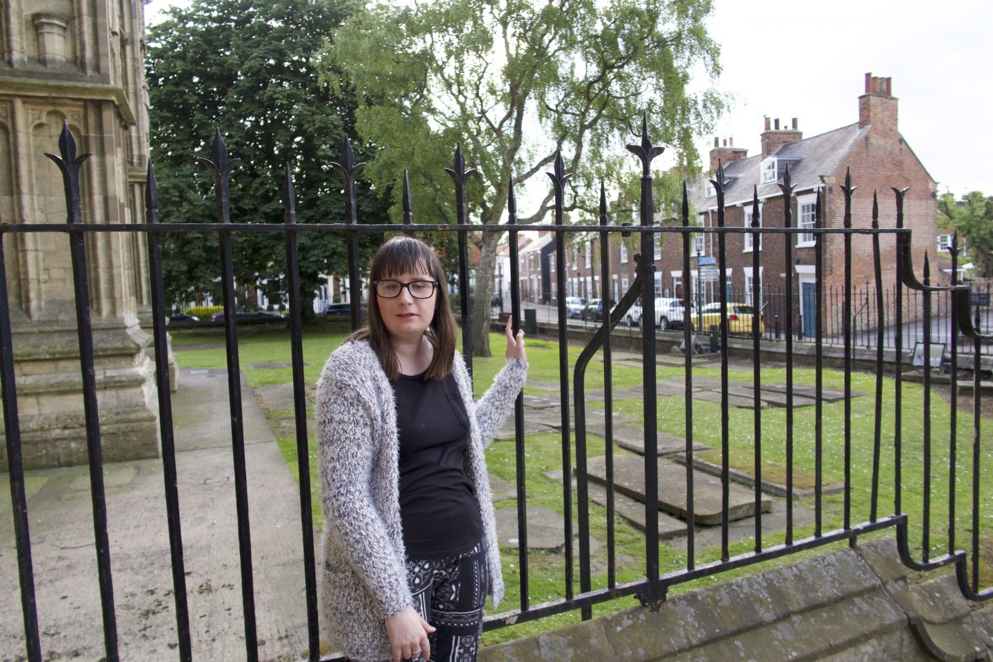 holly stood in front of some railings, there is a Cathedral in the background. She is wearing a black top, black and white trousers and a multi-coloured cardigan
