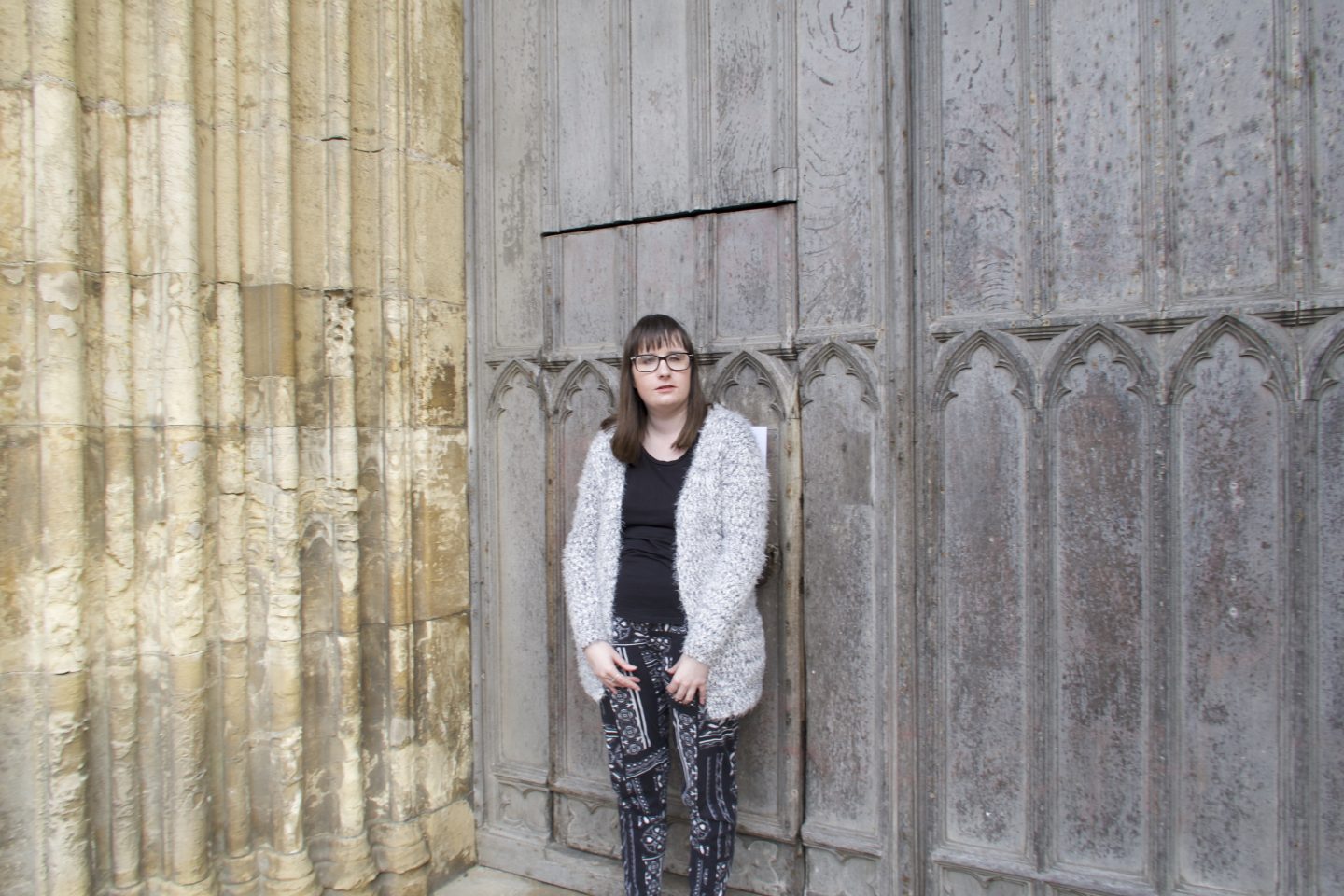 holly stood in front of big metal doors of a Cathedral, she is wearing a black top, black and white trousers and a multi-coloured cardigan