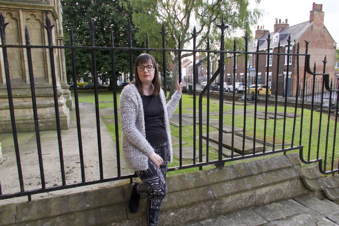 holly stood near some metal bars, her foot is lent against them, there is a Cathedral in the background. She is wearing a multi-coloured cardigan, black top and black and white trousers