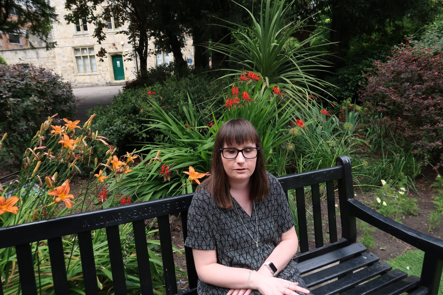 Holly sat on a bench, there are colourful flowery shrubs behind her and a stone building in the background. She is wearing a black and grey dress