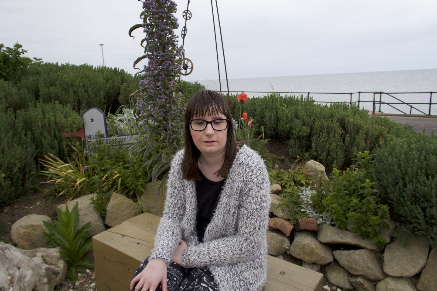 Holly sat on a stone, there is a rockery behind her which has green bushes planted in it and stones surrounding it, the sea is in the distance. She is wearing a multicoloured cardigan and a black top