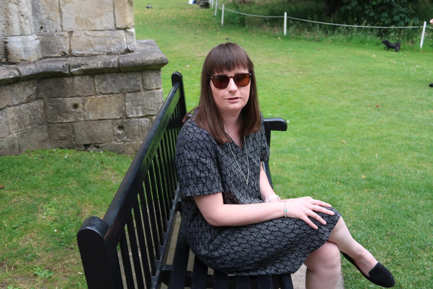Holly sat sideways on a bench, there is a stone building in the background. She is wearing a black and grey dress, black shoes and sunglasses