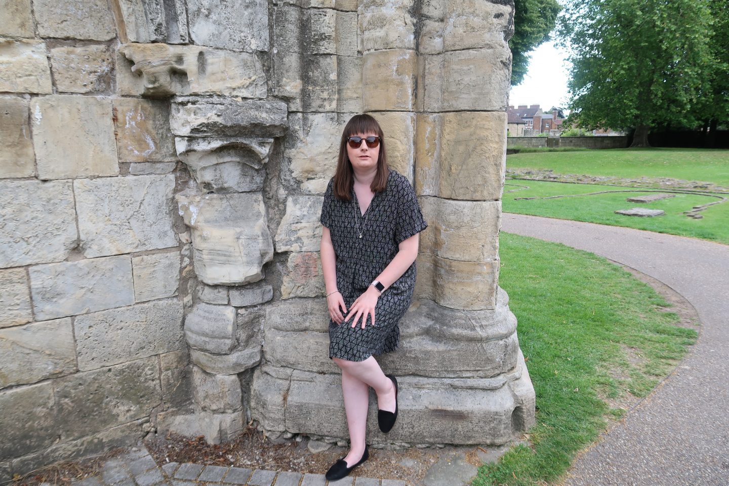 Holly stood in front of a stone wall with her foot up on it and is smiling. She is wearing a black and grey dress and black shoes, she has a silver chain around her neck which has a silver heart on it