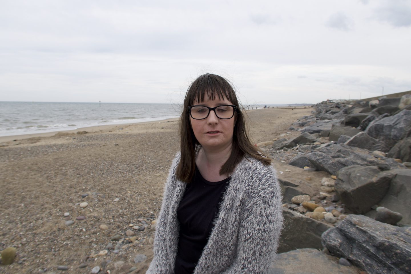 Holly stood on a beach, the sea is in the background and there are rocks to the side, she is wearing a black top and a woollen multicoloured cardigan