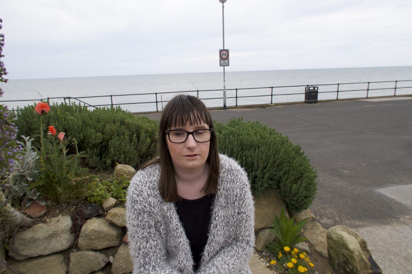 Holly sat on a stone, there is a rockery, green bushes and red poppies behind her and the sea is in the distance. She is wearing a multicoloured cardigan, she is looking to the right