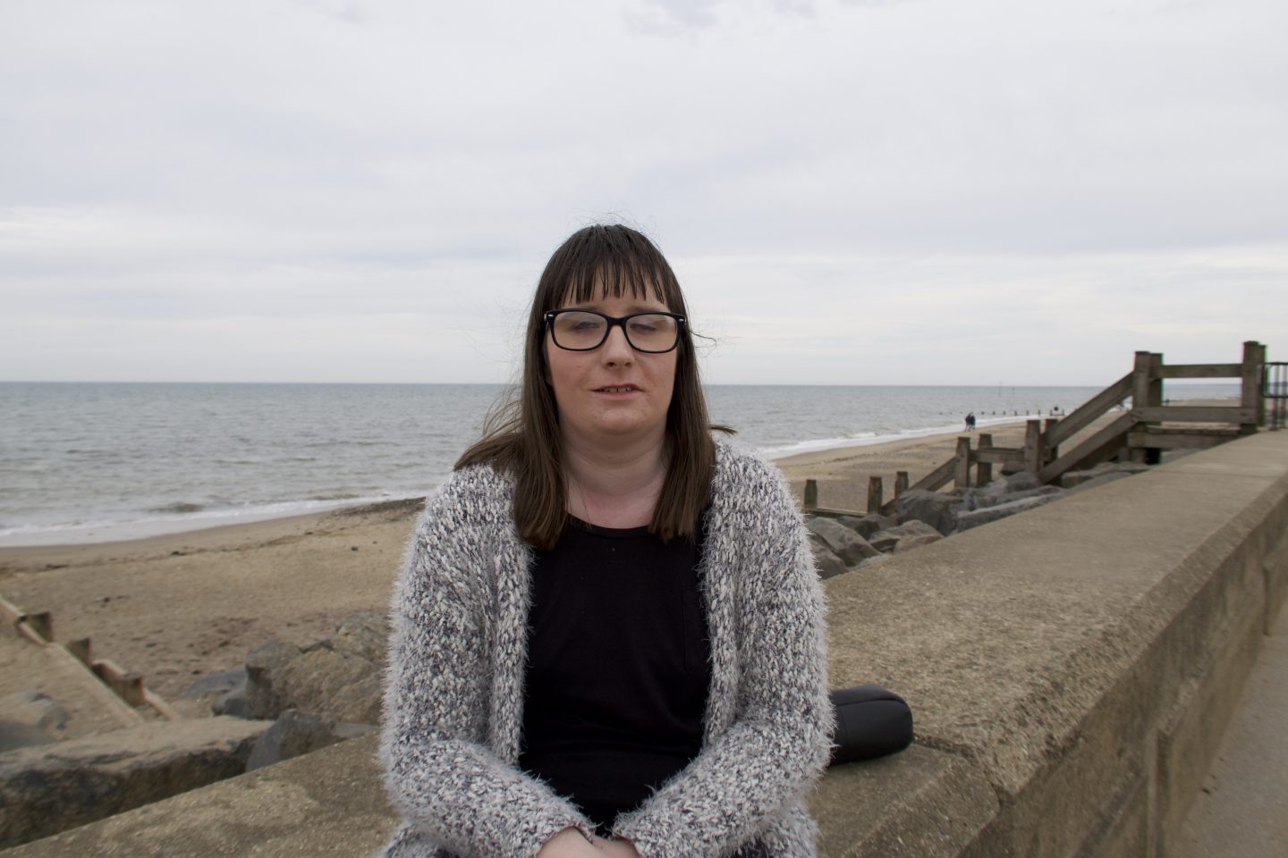 Holly sat on a wall, the beach and sea are in the distance behind her. She is wearing a black top and a multicoloured cardigan