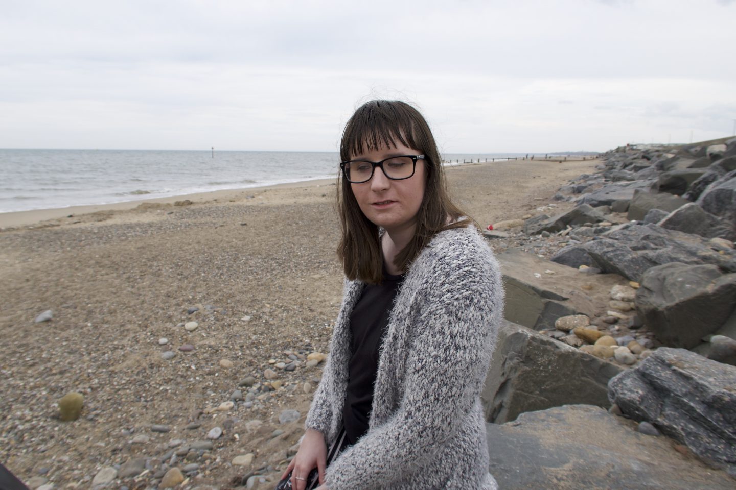 Holly stood on a beach looking down, the sea is in the background and there are rocks to the side. She is wearing a multicoloured cardigan and a black top