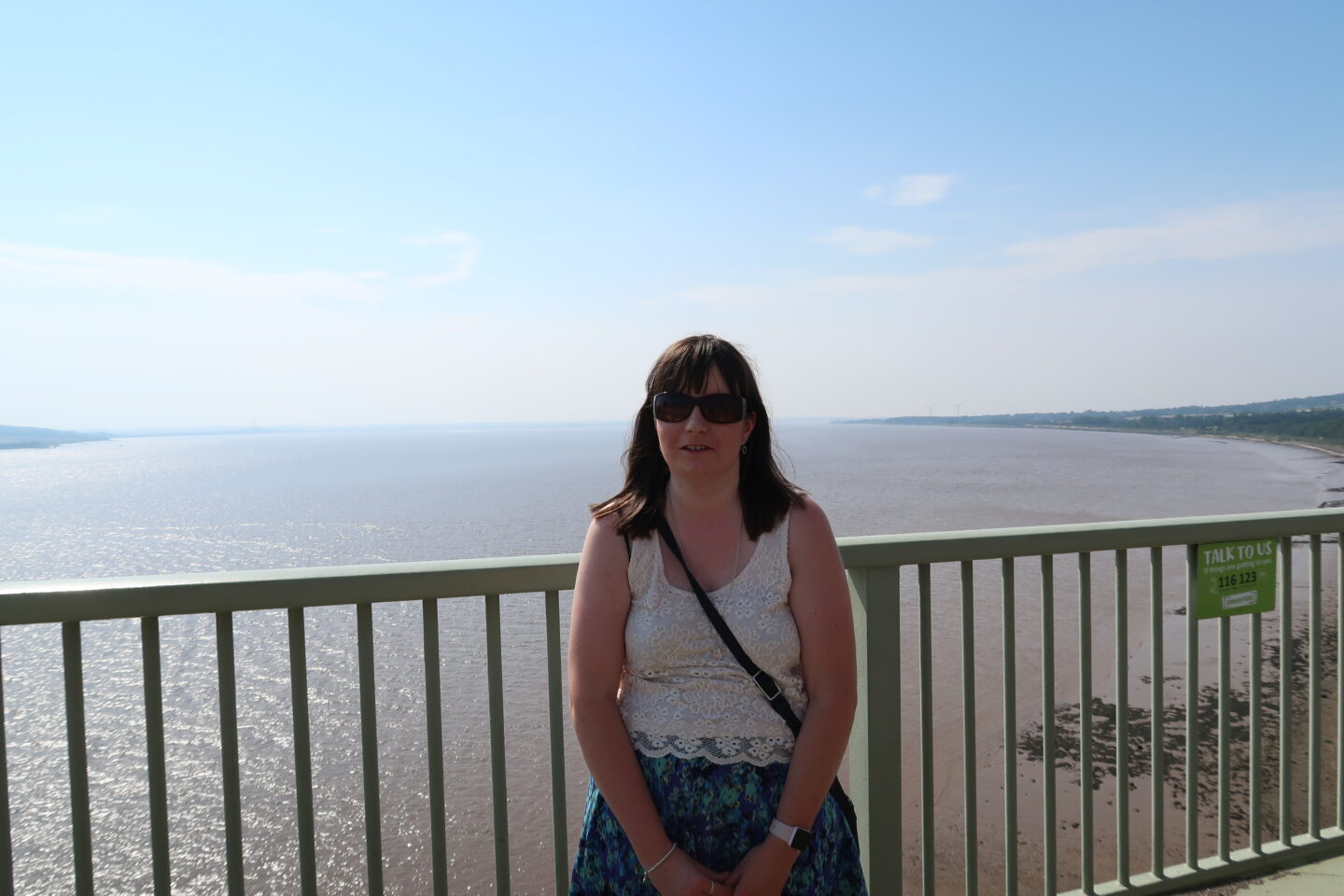 A close-up photo of Holly stood on a high bridge, the sea and green fields are in the background. She is wearing a cream coloured lace top, a multicoloured flowered skirt, and sunglasses