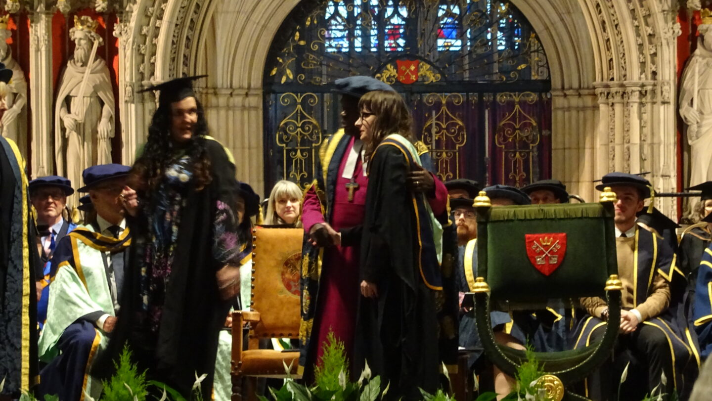 Holly receiving her graduation certificate at York Minster, people can be seen on a stage in the background. She is wearing a black graduation gown and a gold and blue sash