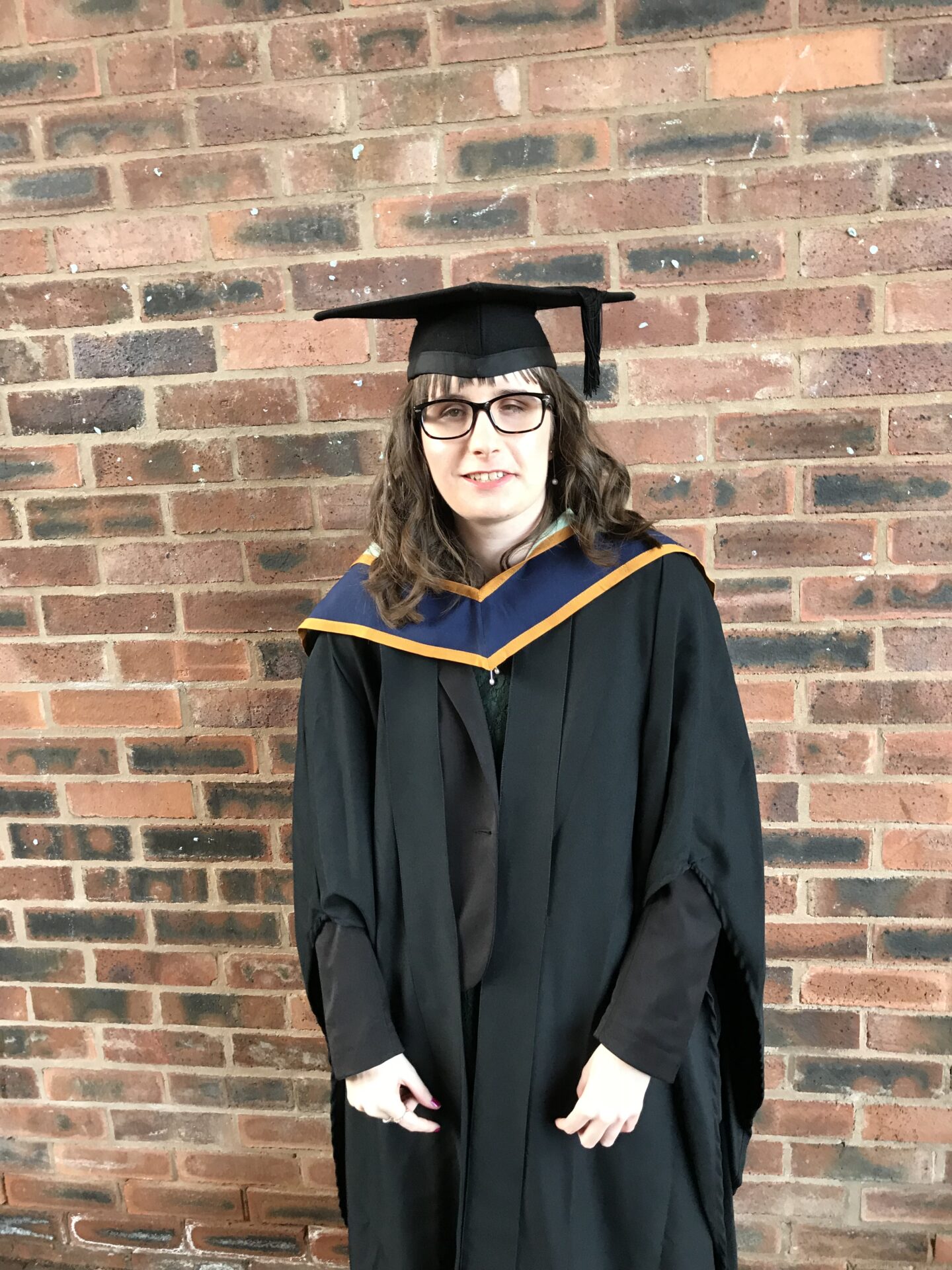 Holly stood in front of a wall, she is wearing a black mortarboard, a black graduation gown and a gold and blue sash