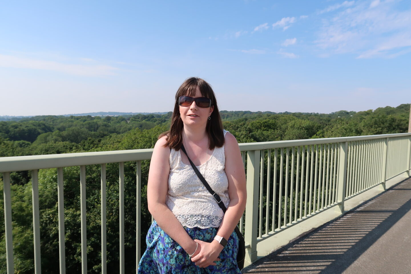 Holly stood on a high bridge in front of a large railing, there is a mass of green trees in the background and a lovely blue sky can be seen. She is wearing a cream coloured lace top, a multicoloured flowered skirt and sunglasses