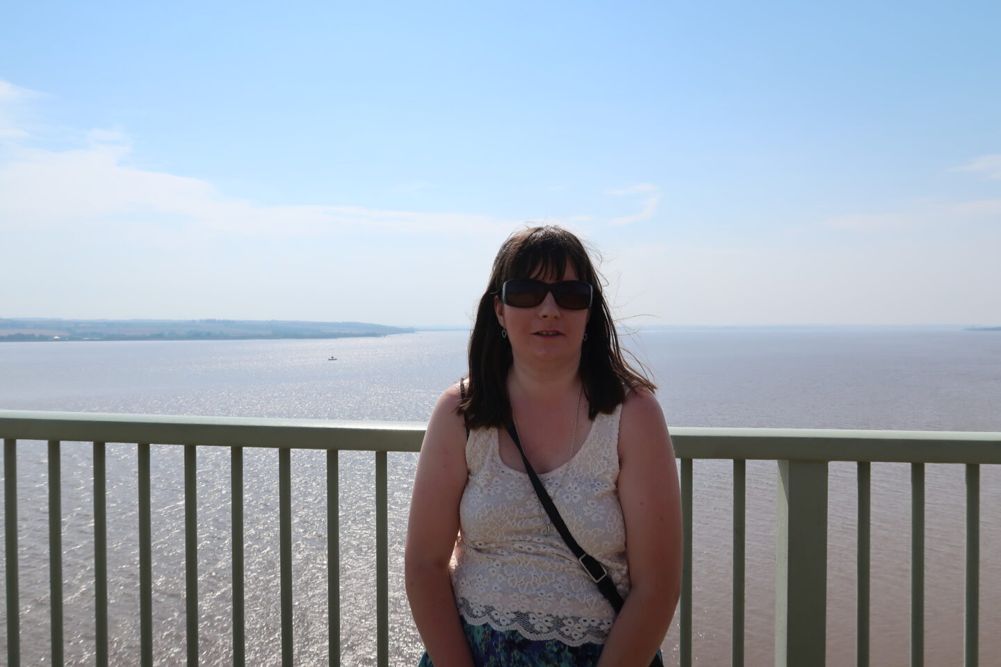 Holly stood on a high bridge, She is wearing a cream coloured lace top, a multicoloured flowered skirt and sunglasses and is smiling. The sea can be seen in the distance