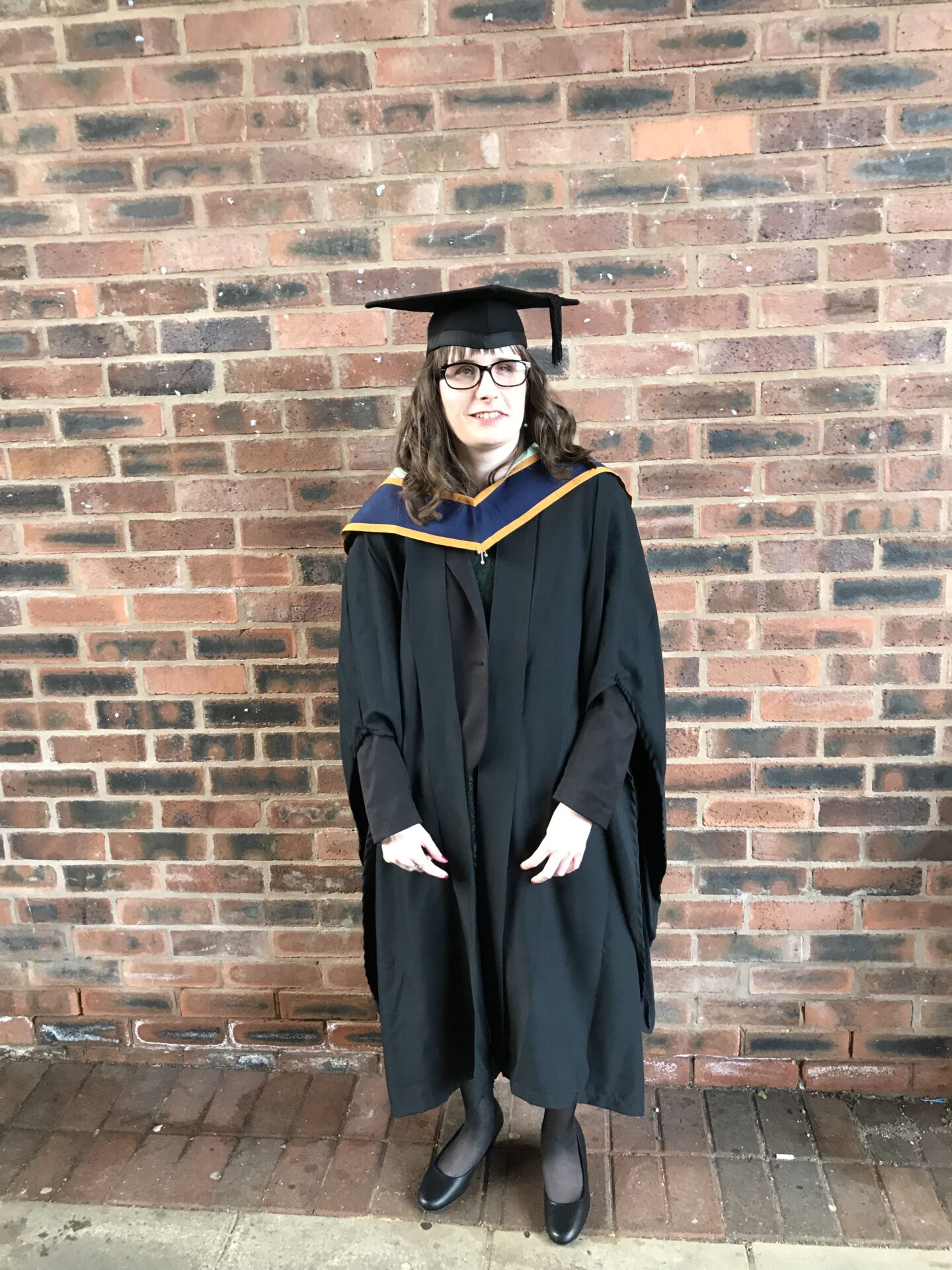A portrait photo of Holly stood in front of a wall, she is wearing a black graduation gown, a blue and gold sash, black tights and black shoes