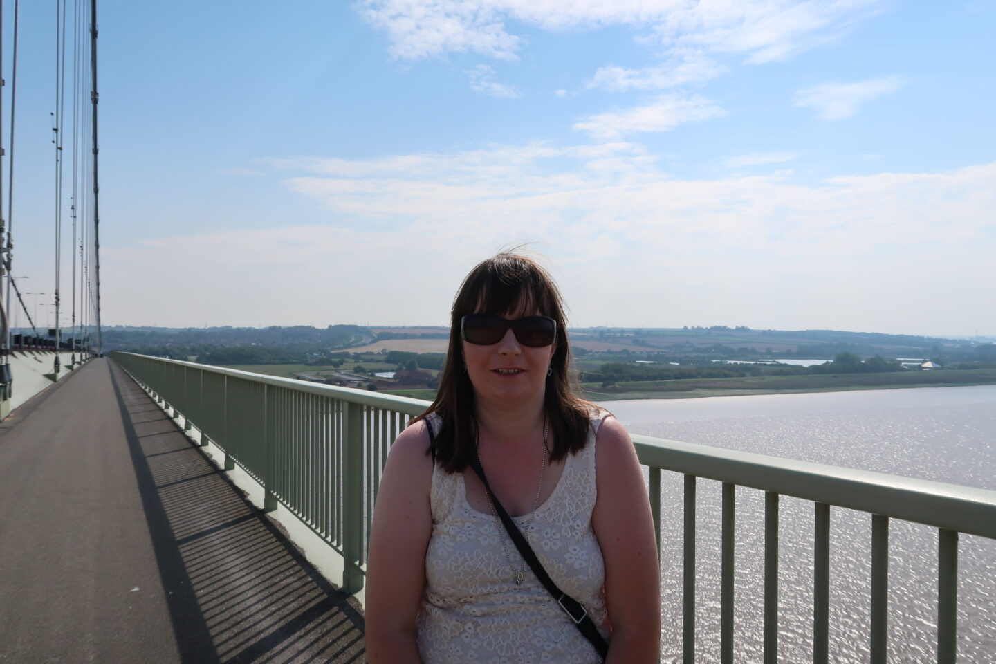 Holly stood smiling on a high bridge, she is wearing a cream coloured lace top, a flowered skirt and sunglasses. A long path, the sea and green land can be seen in the distance