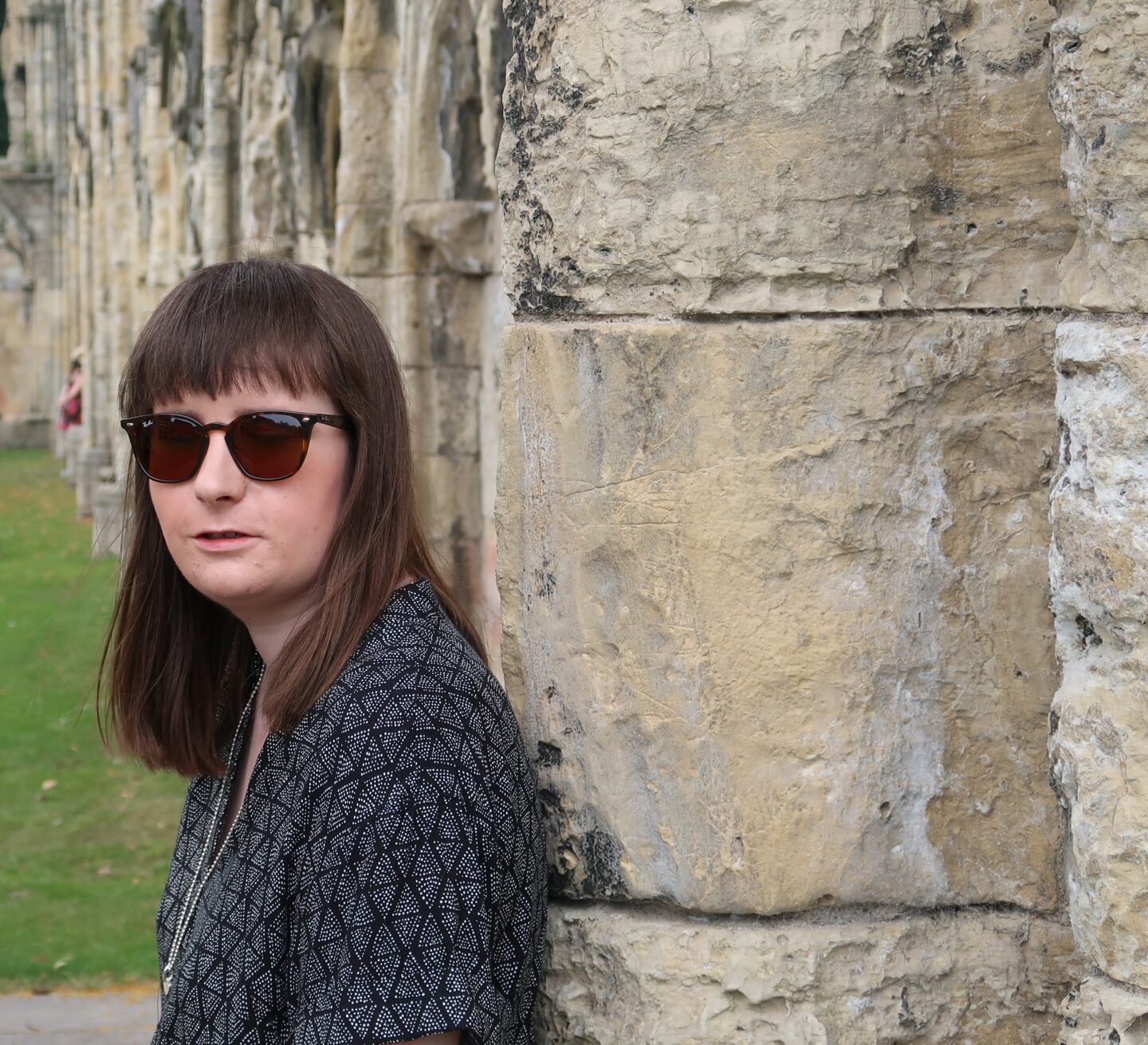 A close up photo of Holly leant against a stone wall. She's wearing a black and grey dress and sunglasses