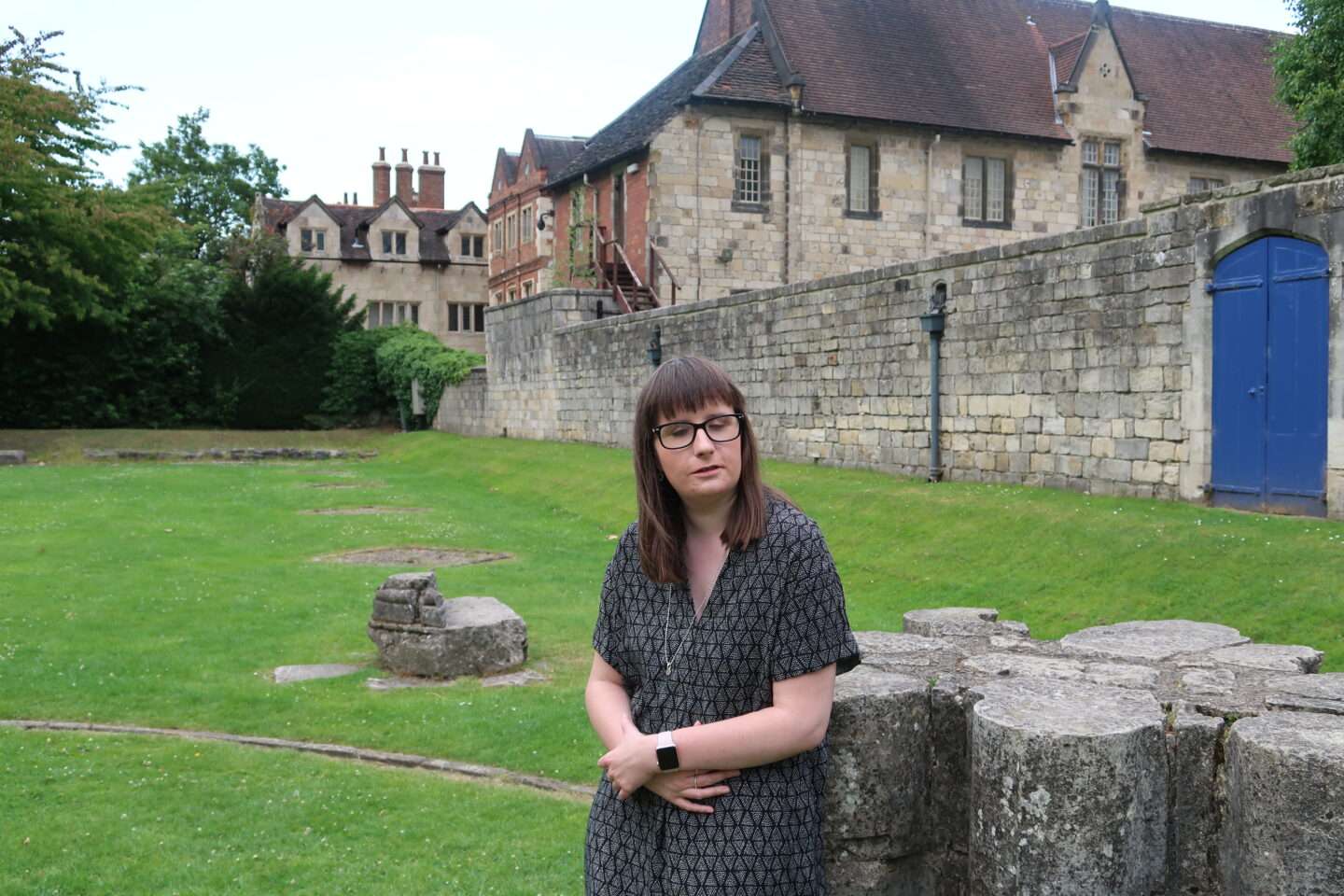 Holly leant against a small stone monument in front of some very old stone buildings, she is wearing a black and grey dress