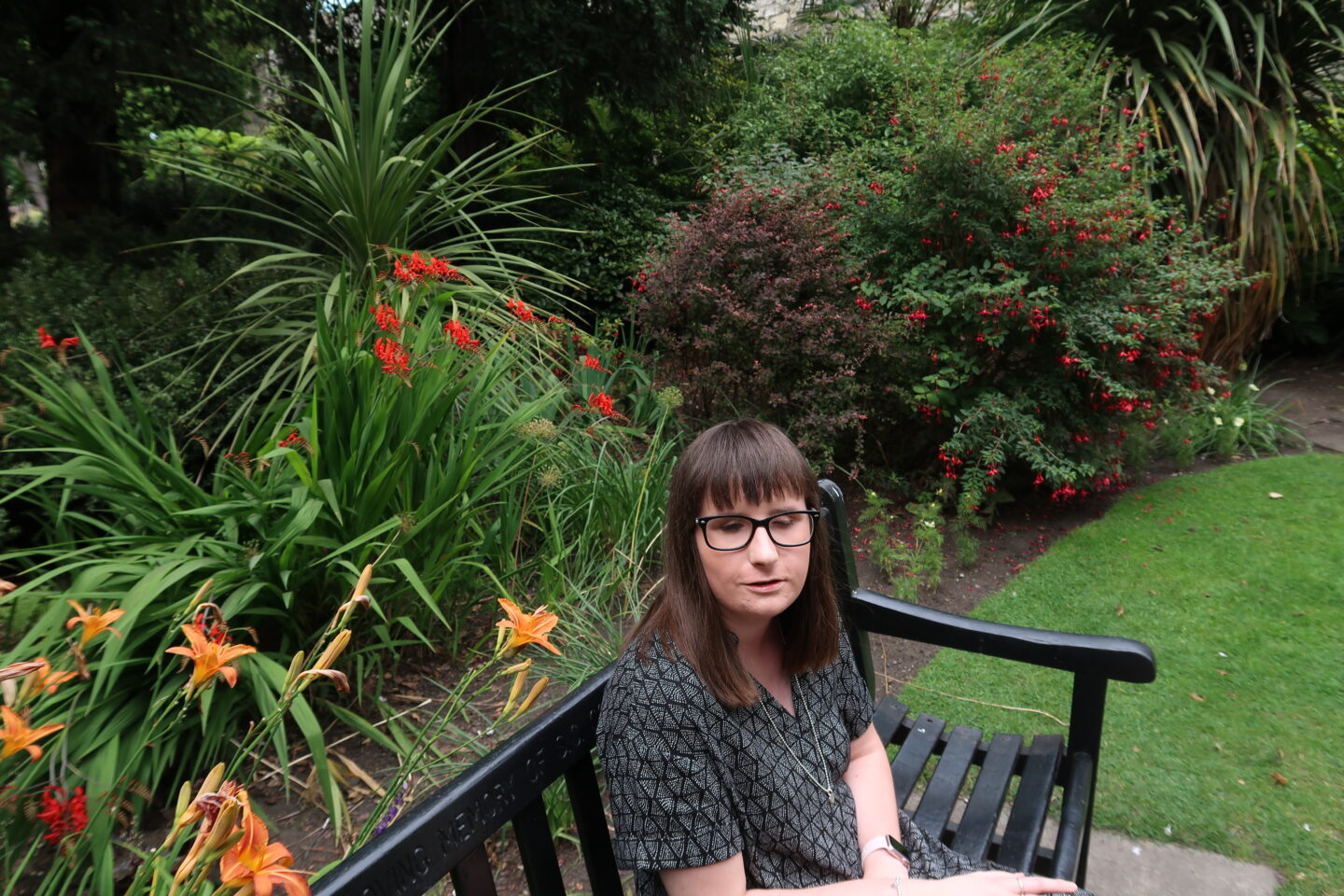 Holly sat on a bench, there are colourful flowers behind her. She is wearing a black and grey dress