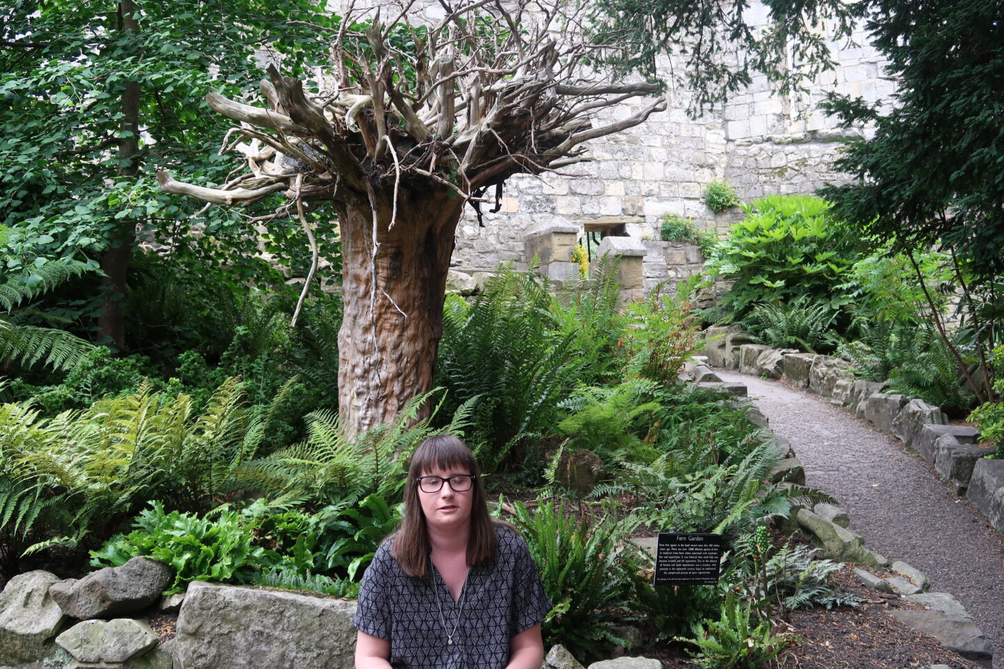 Holly sat on a stone, there are green trees and a stone wall behind her. She is wearing a black and grey dress
