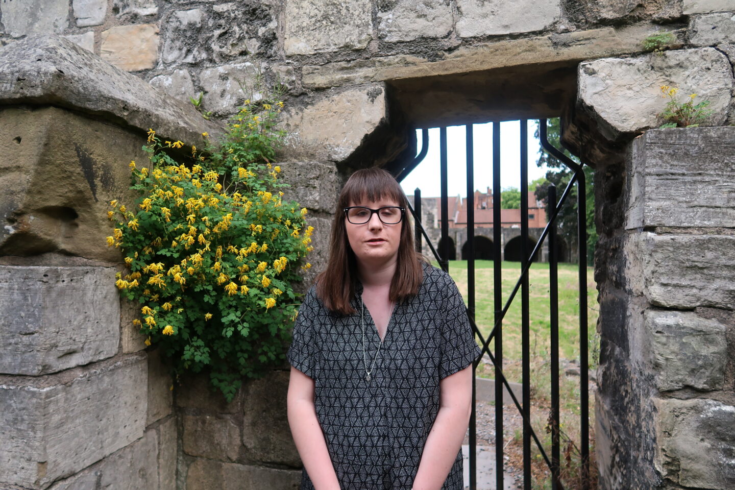 Holly stood in front of a black iron gate, there is a stone wall and a green bush to the right. She is wearing a black and grey dress