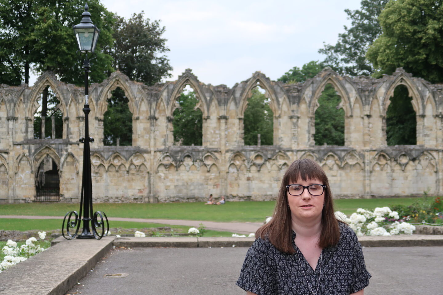 Holly stood in front of a stone wall which has trees showing through the gaps of some arches within it, a very old black tall lamp can be seen in the background. She is wearing a black and grey dress