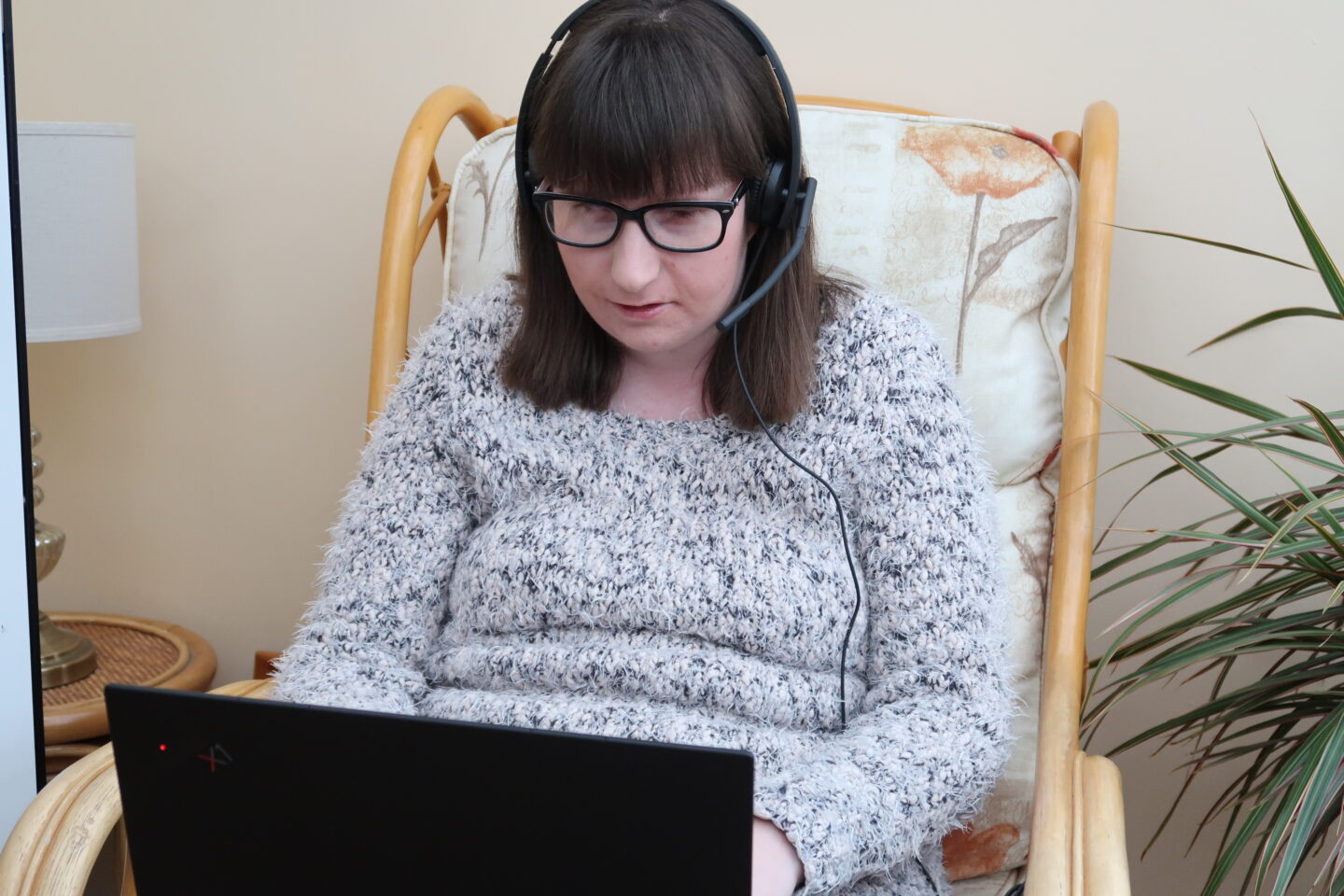 Holly sat in a chair in a conservatory, she has a headset on and has a laptop on her knee. She is wearing a cream and grey fluffy jumper and black leggings
