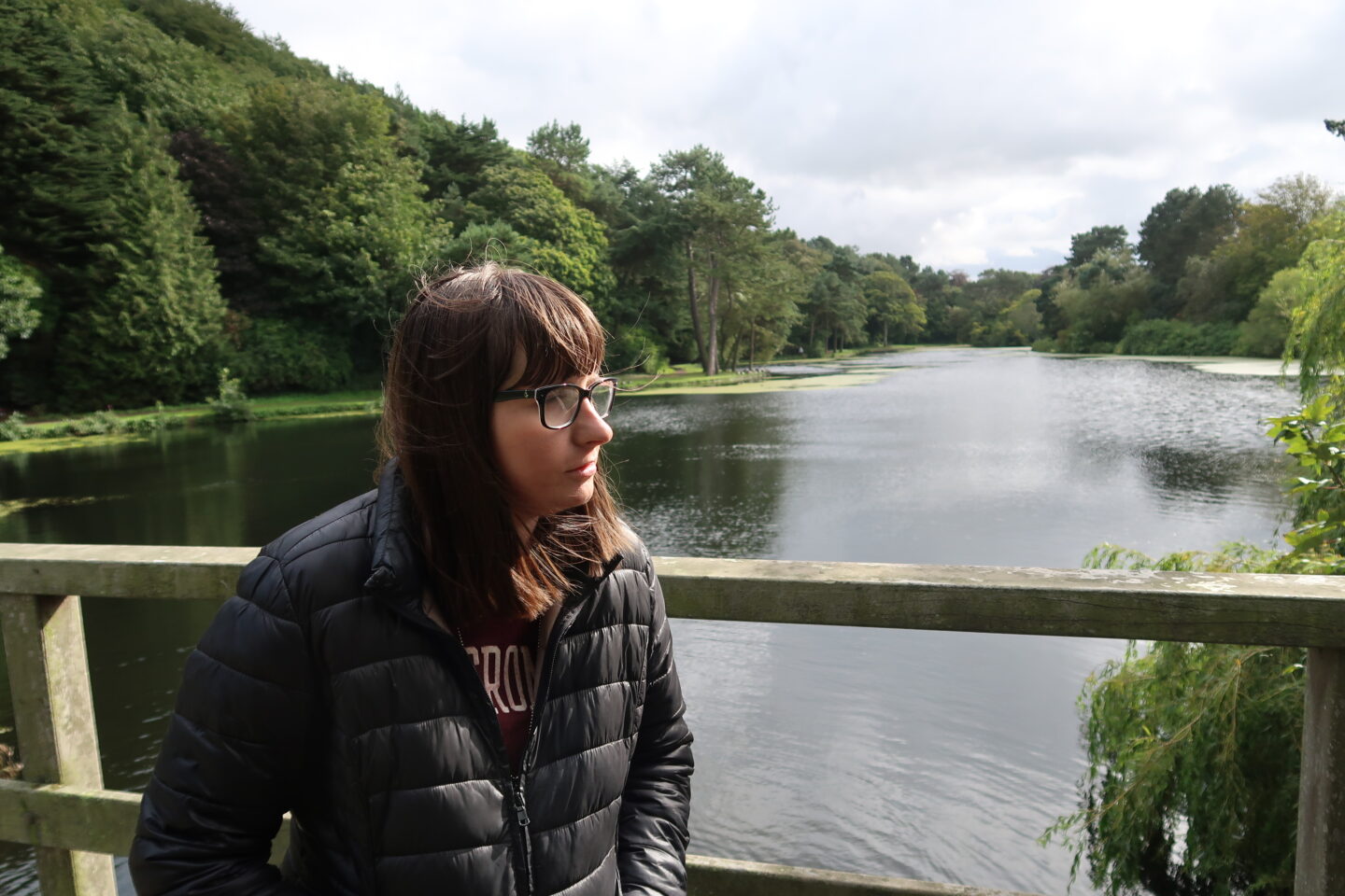 Holly stood against a wooden fence looking to the side, there is a lake and green trees in the distance. She is wearing a burgundy top and a black jacket